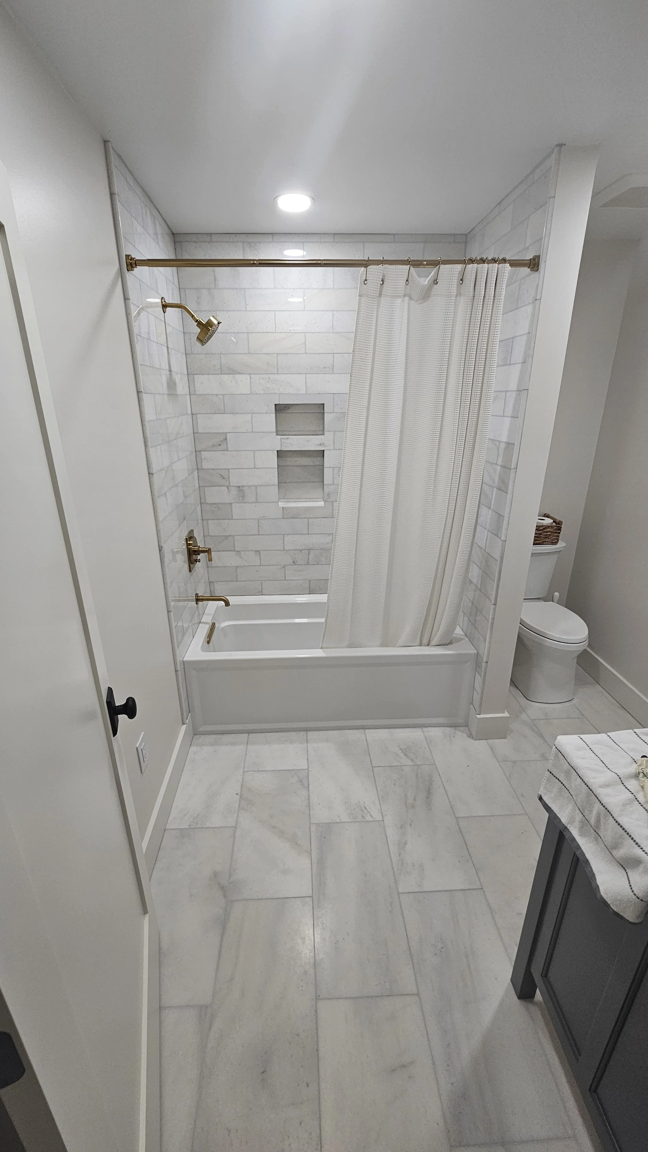 Bathroom with white tiled walls, a bathtub with a shower curtain, and a toilet. The floor is tiled with gray and white stone-like tiles.
