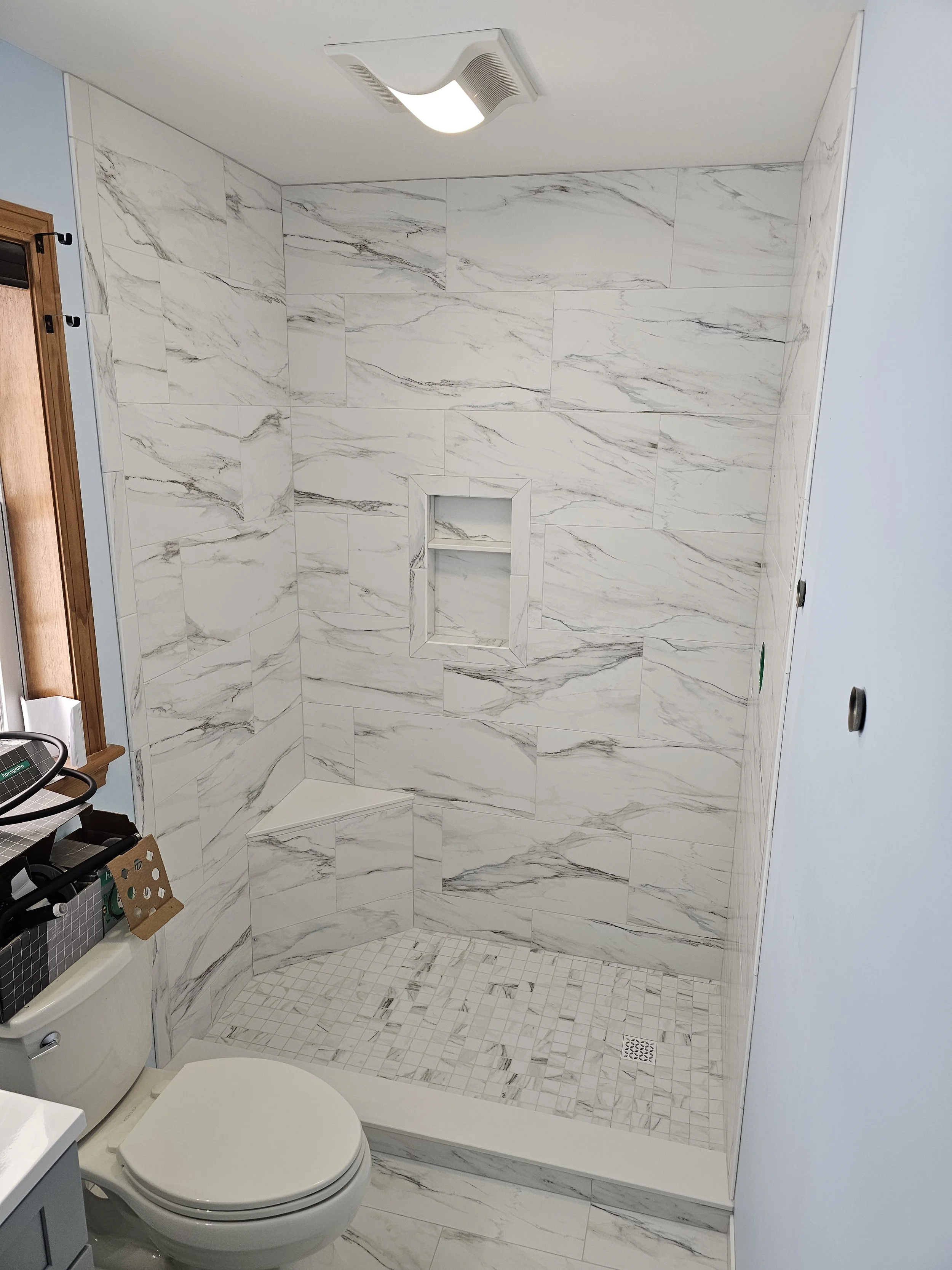 Small bathroom with a corner shower that has marble-like tiled walls and a built-in shower niche. The floor has small square tiles, and there's a toilet and part of a vanity visible. The ceiling has a vent and a light fixture.