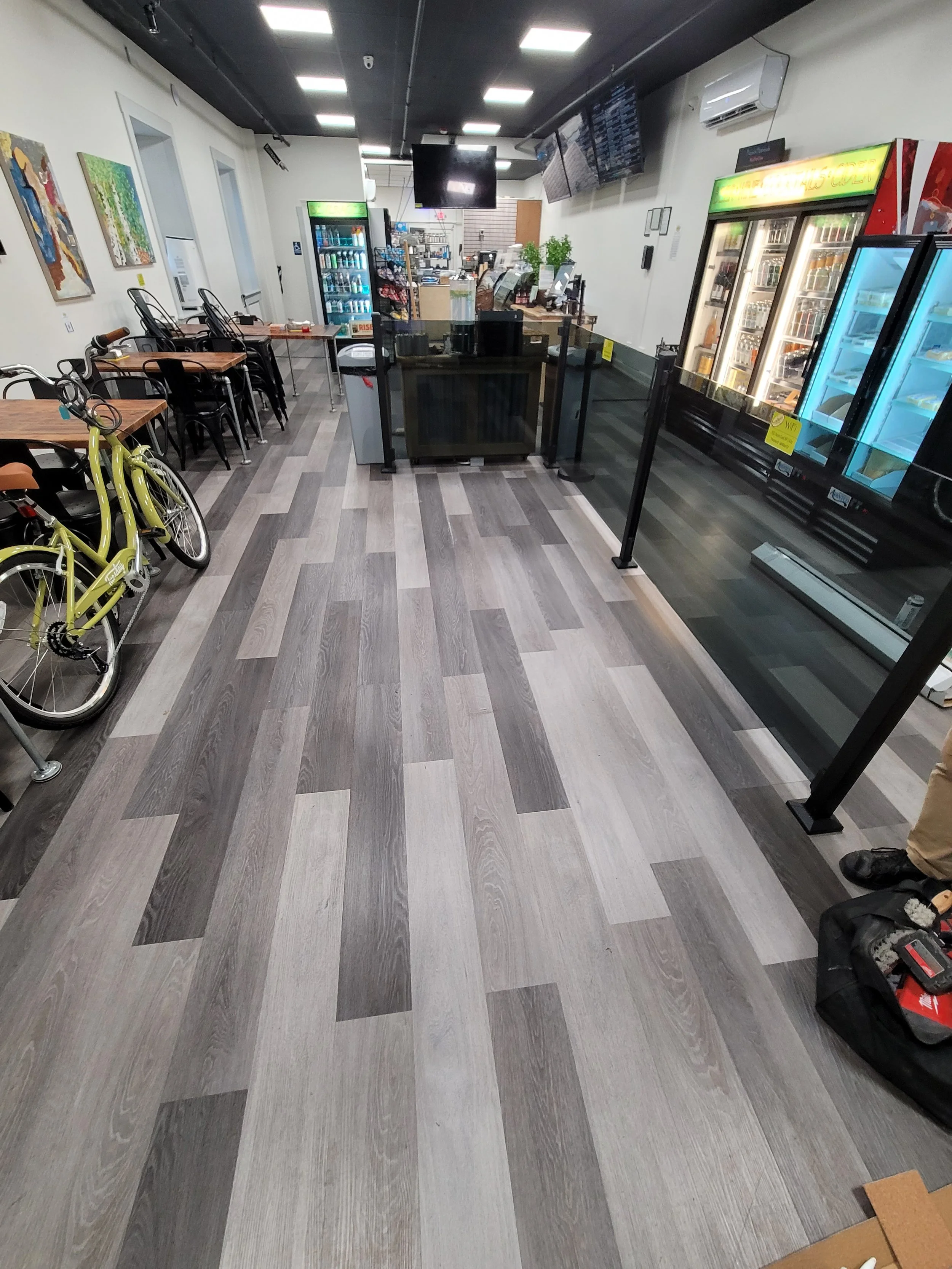Inside a modern coffee shop or convenience store with wood-patterned flooring, a bicycle on the left, and refrigerated beverage coolers on the right. The background shows counter areas, snack racks, and the overall layout.