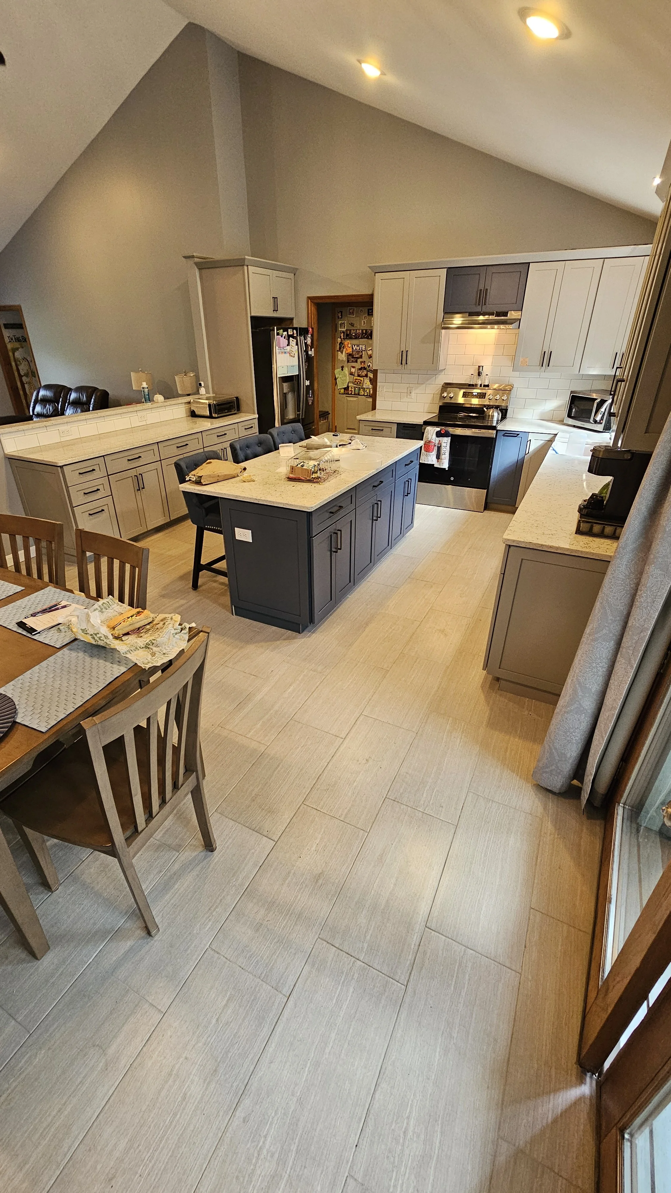 Interior view of a kitchen with a large central island, beige cabinets, stainless steel stove, refrigerator, and microwave, and a dining table with chairs.