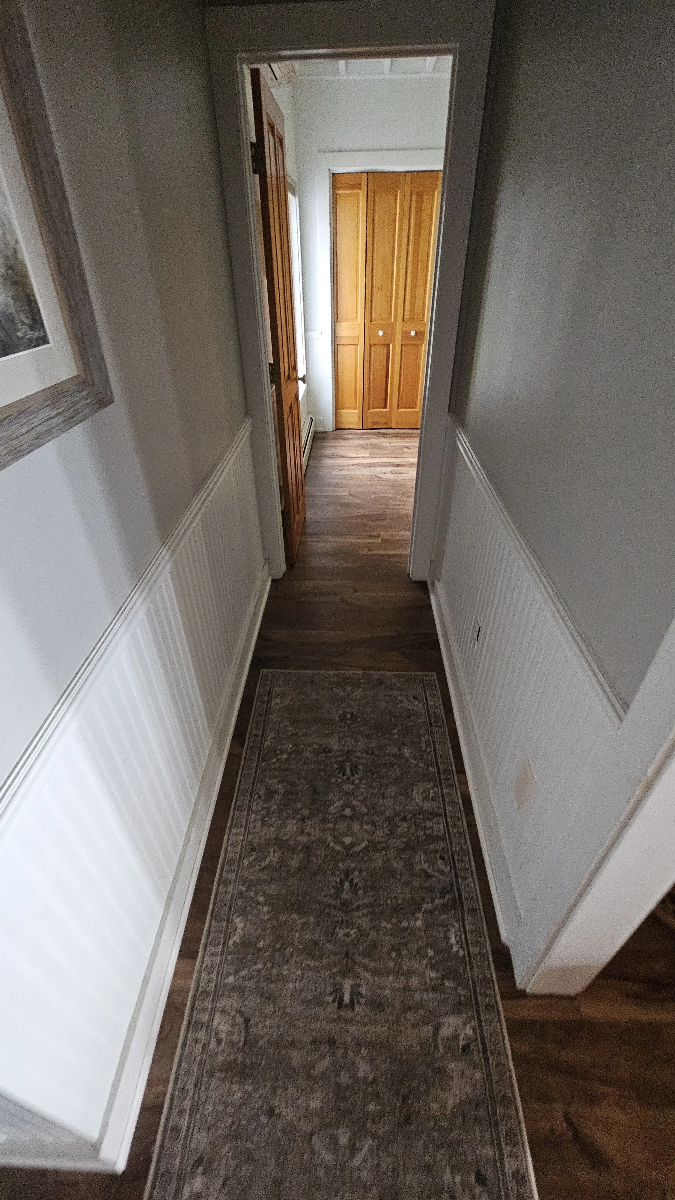 A narrow hallway with hardwood flooring, white walls with wainscoting, a runner rug, and a doorway at the end leading to a room with wooden doors.