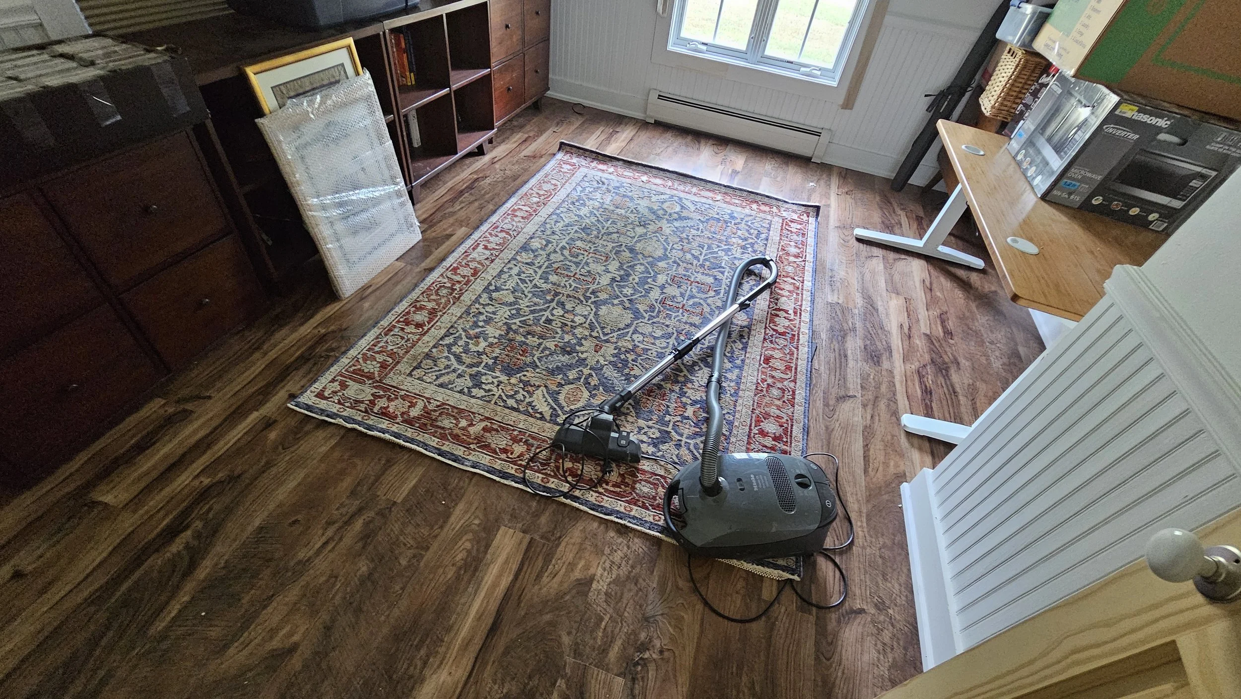 A room with hardwood floors, a patterned area rug, a vacuum cleaner with a hose, a vacuum attachment, and a vacuum cord. There are bookshelves, a framed picture, and a window with a view outside. A wooden table holds a microwave oven and some boxes.