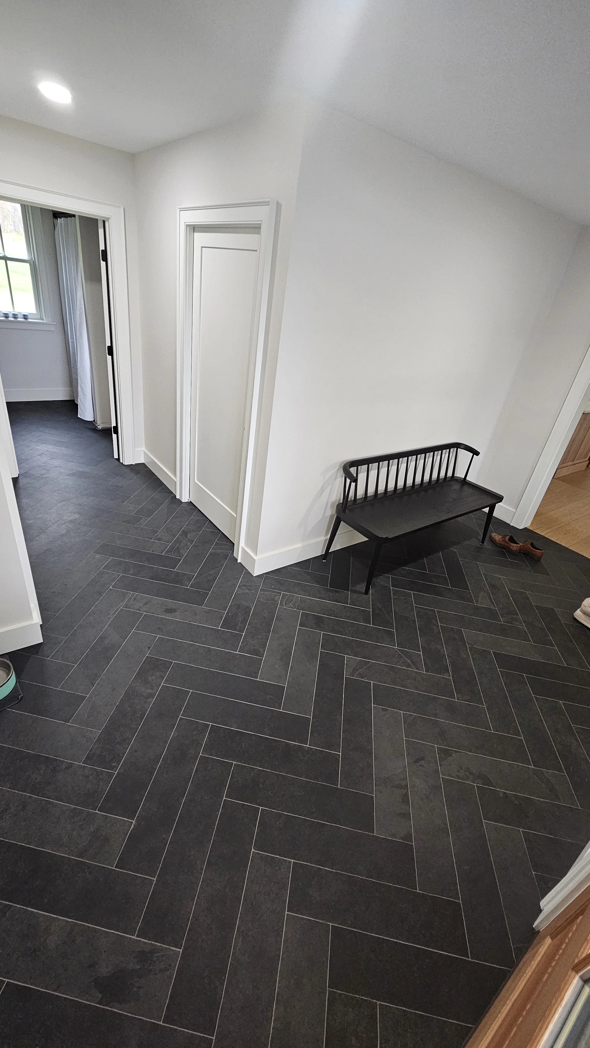 Interior view of a house with dark tiled flooring, white walls, a black bench, and a pair of shoes on the floor. There are doorways leading to other rooms.