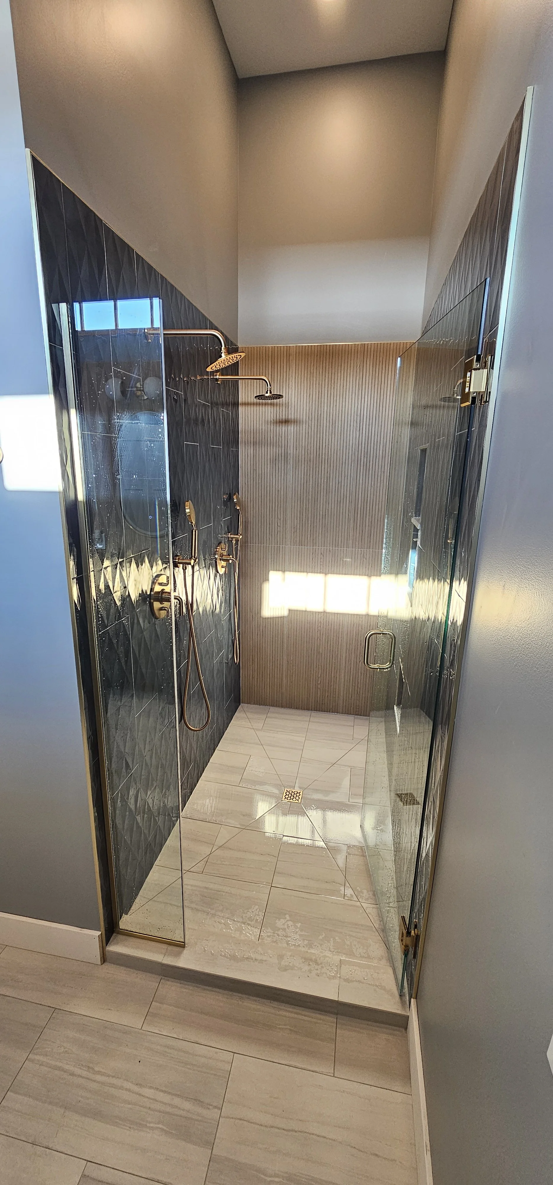 Modern walk-in shower with black diamond-patterned tiles on one wall, beige wood-textured tiles on the other walls, and two gold shower fixtures with handheld and overhead showerheads. The shower has glass doors and a window that lets in natural light.