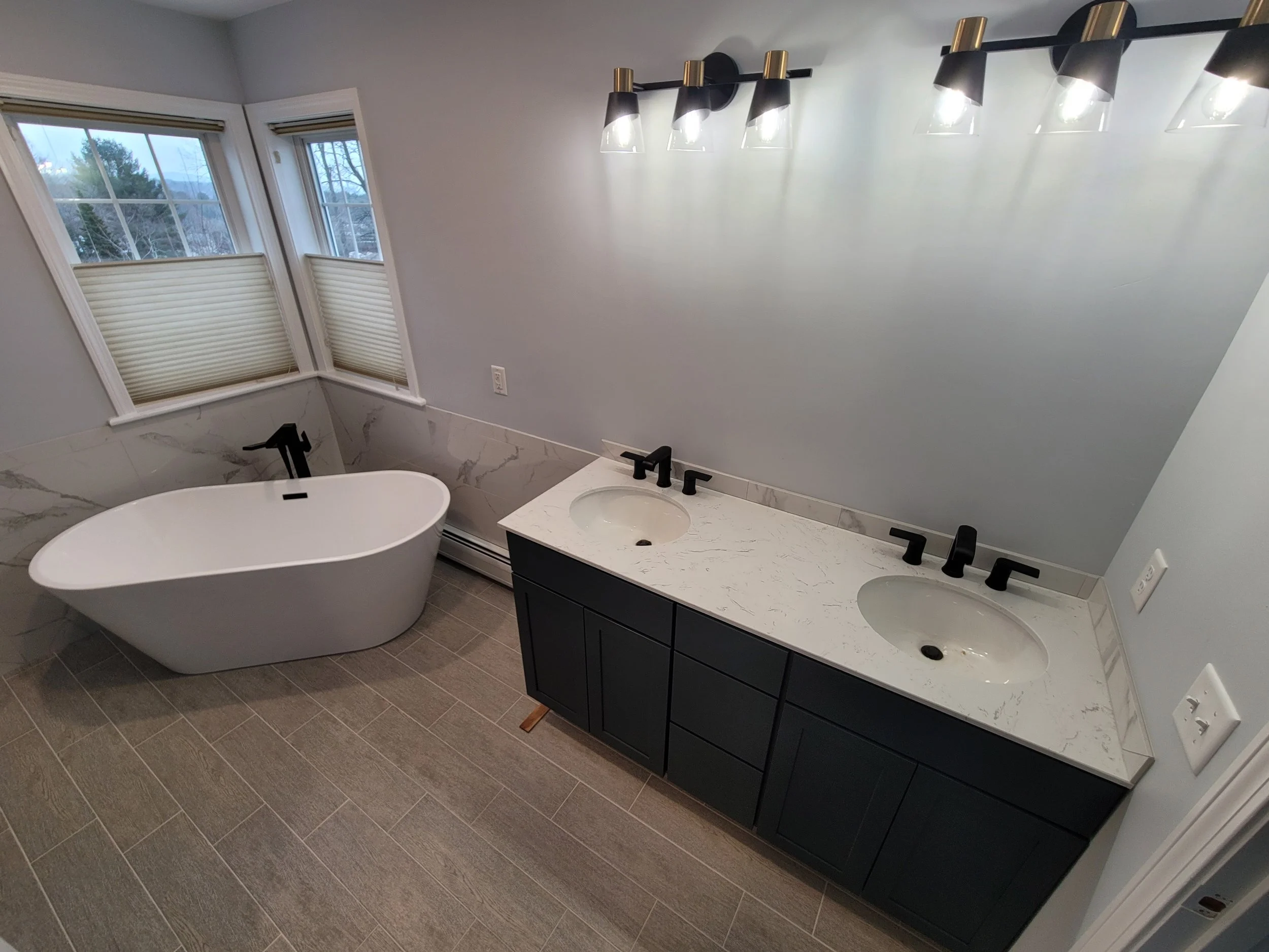 A modern bathroom with a freestanding bathtub, a double sink vanity with black cabinets and marble countertop, and a large window with white blinds. There are four wall-mounted light fixtures above the mirror.