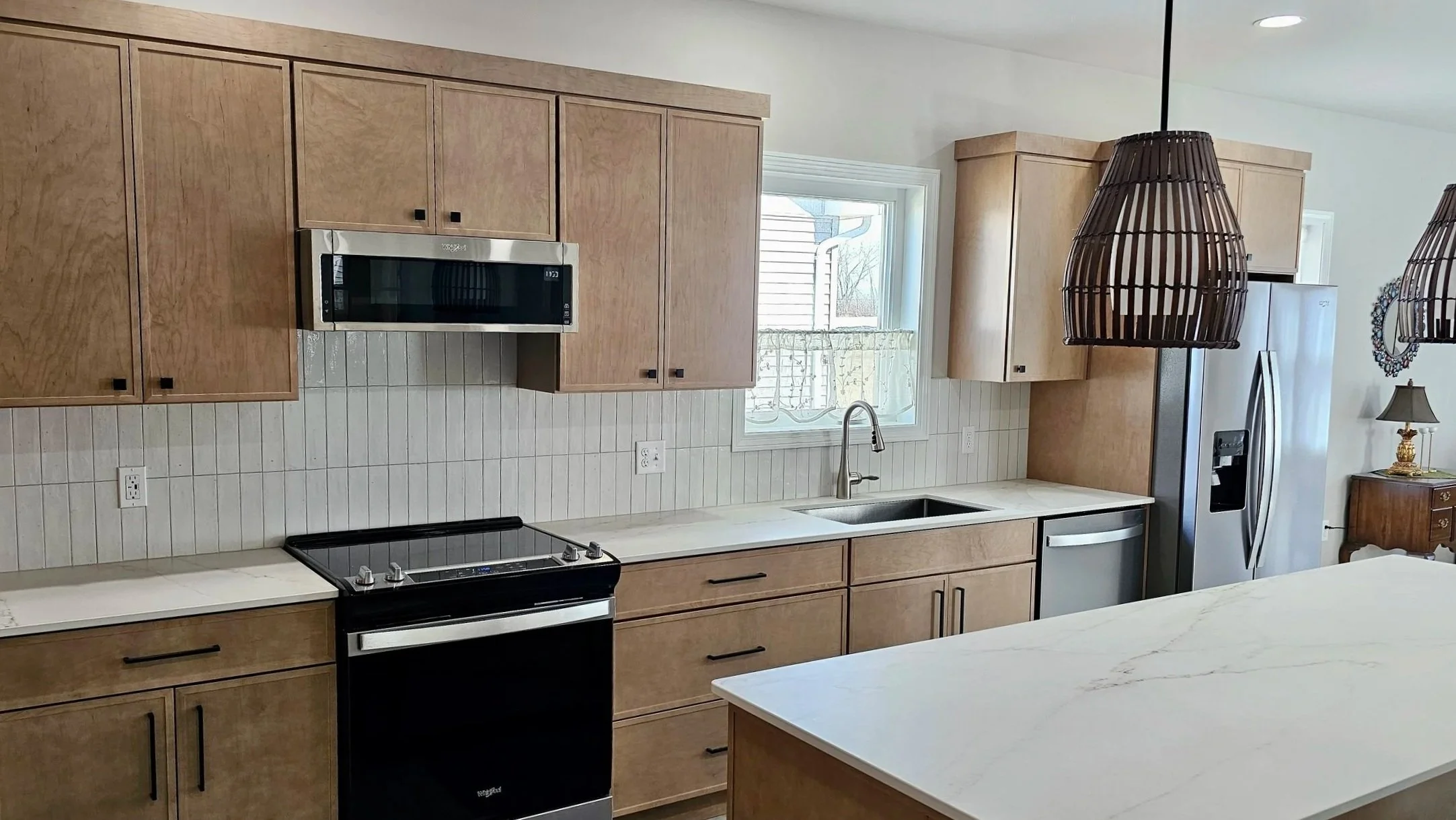 Kitchen with upper and lower wooden cabinets, a window above the sink, a stainless steel microwave, a black stove, and white countertops. A wicker pendant light hangs over the counter, and a stainless steel refrigerator is in the corner.