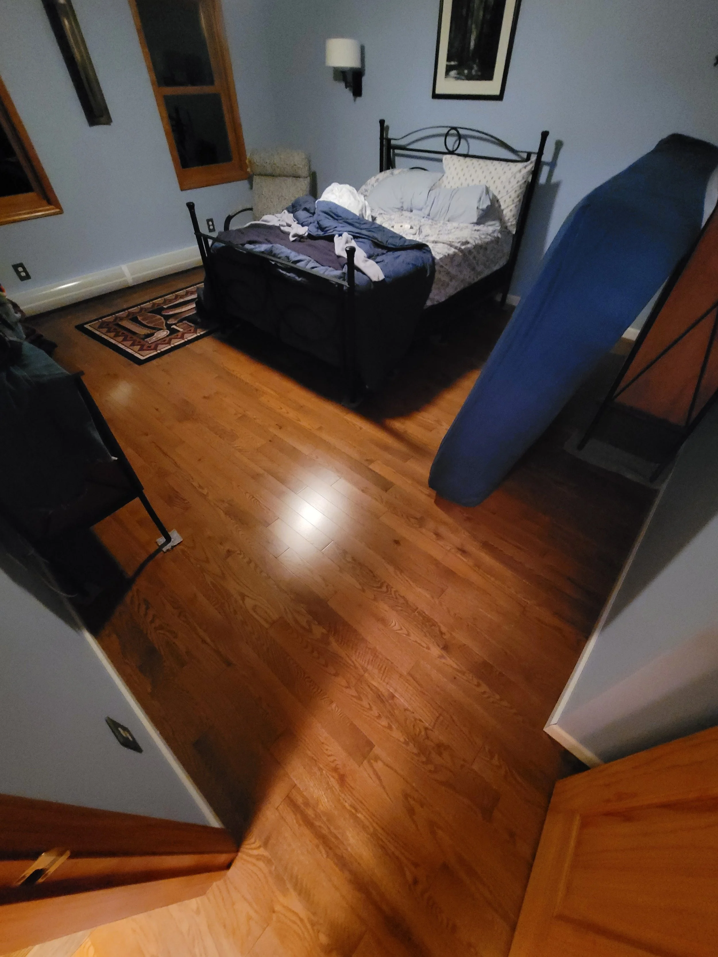 A bedroom with a black metal bed frame, unmade bedding, a small hanging lamp, framed art on the wall, hardwood floor, and an ironing board leaning against the wall.