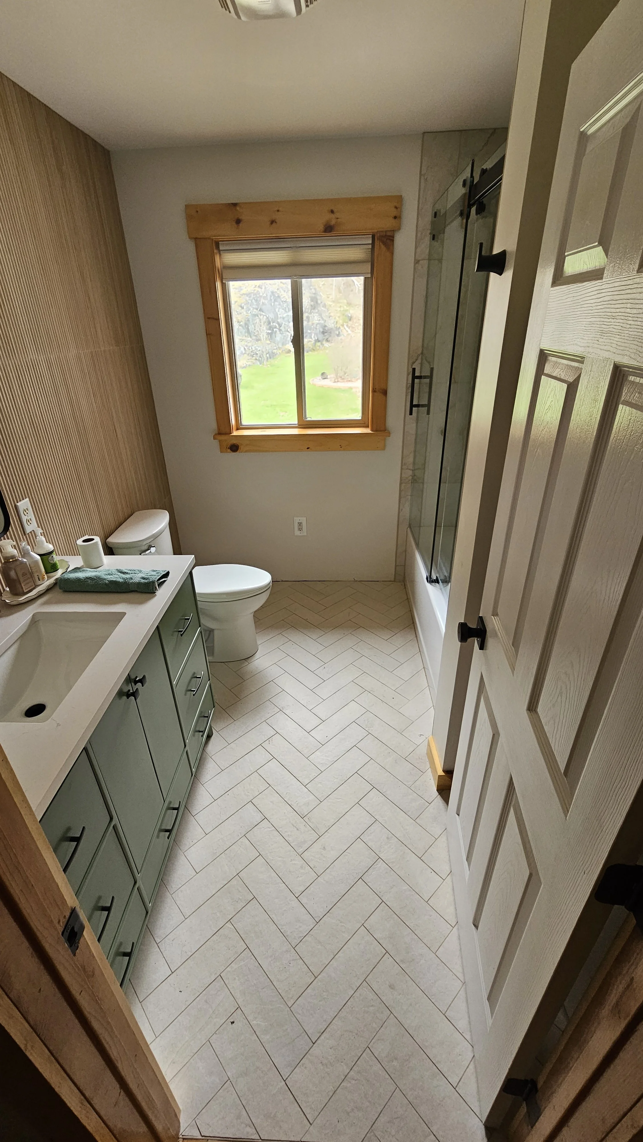 A small bathroom with a window showing a grassy yard outdoors. The bathroom has a white vanity with a green cabinet, a toilet, and a glass shower door. The floor is tiled in a herringbone pattern, and there's a wooden window frame.