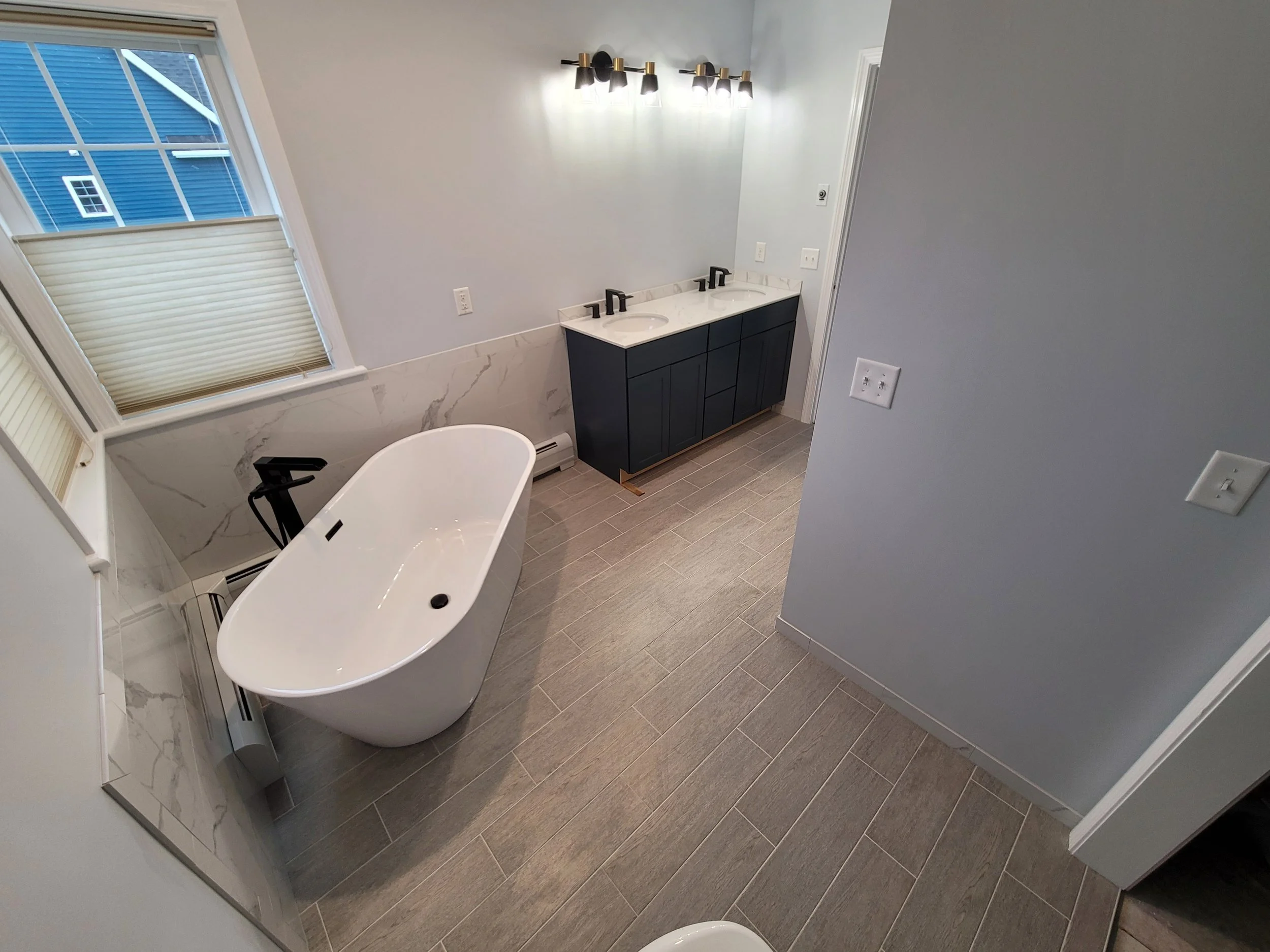 Modern bathroom with double vanity, black cabinets, marble countertops, and black fixtures. There is a bathtub, a window with blinds, and a wall-mounted light fixture.