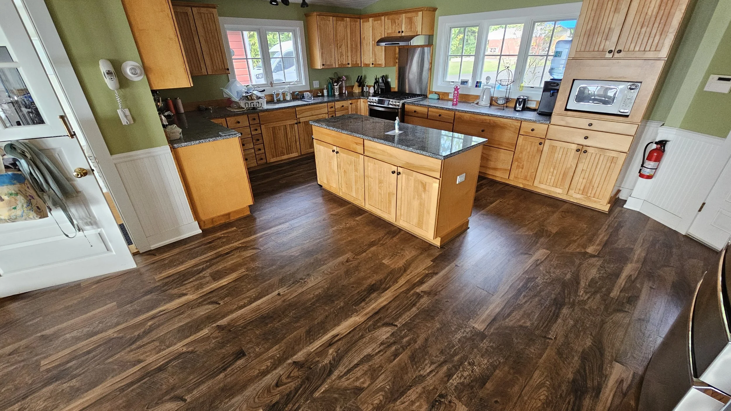 A kitchen with wooden cabinets, granite countertops, and dark wooden flooring. There are windows above the sink and counter, and various kitchen appliances on the counters. An island is in the center of the room, and a fire extinguisher is mounted on
