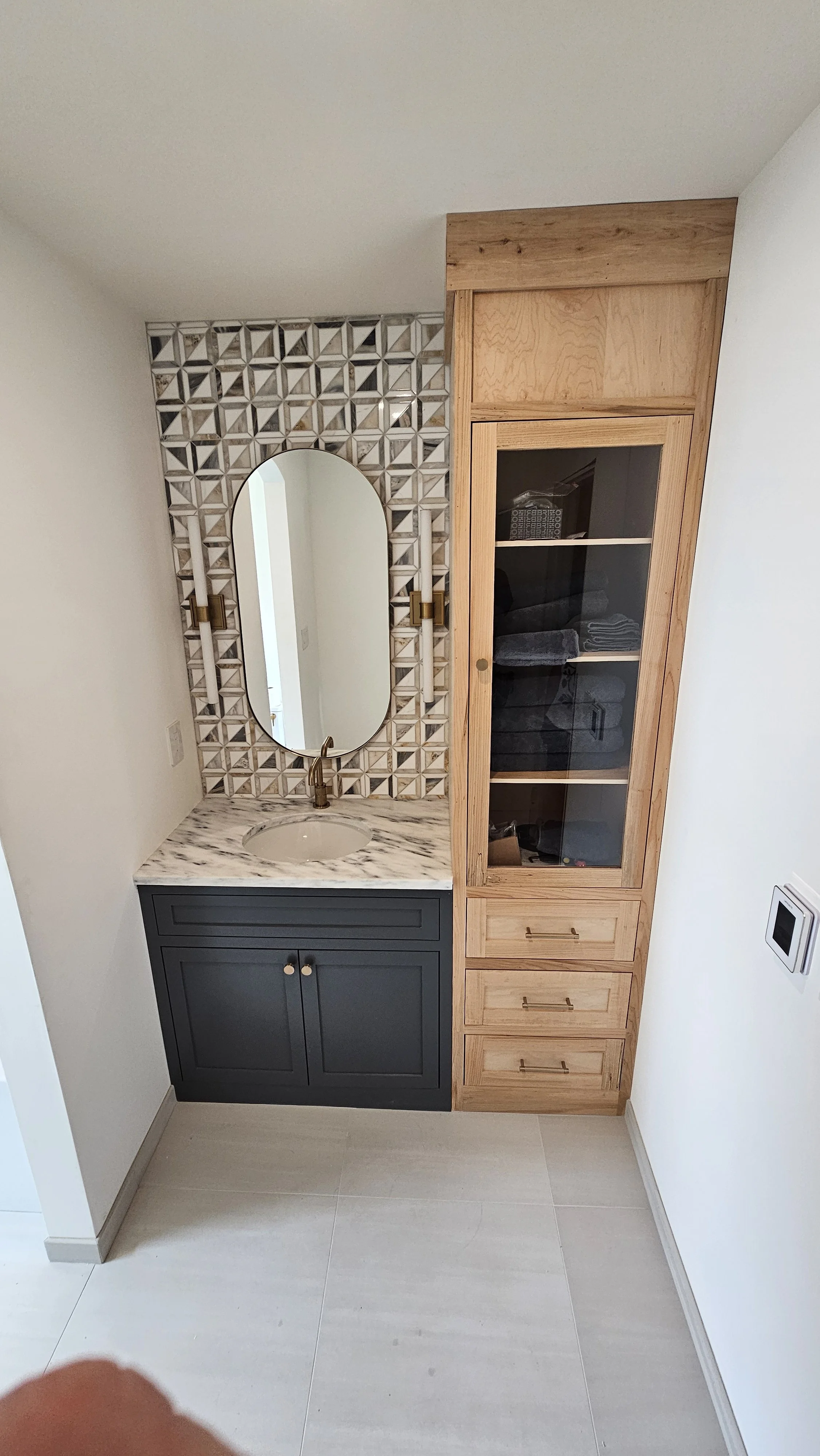 A bathroom vanity with a marble countertop, integrated sink, and a large mirror above, flanked by wall-mounted light fixtures. To the right, a tall wooden cabinet with glass doors and drawers, all set against white walls and light-colored flooring.