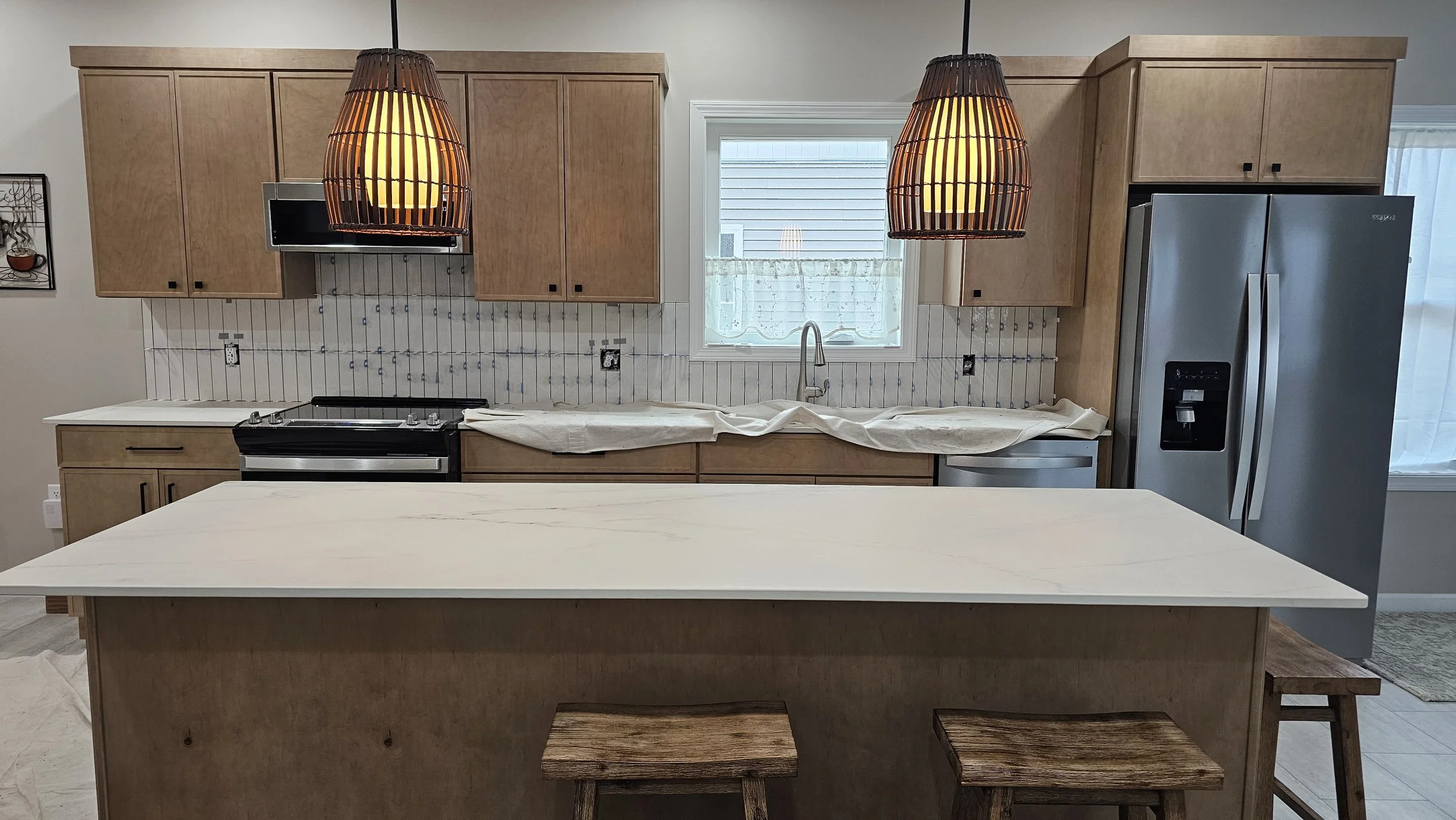 A kitchen in the process of being renovated, with cabinets, a dishwasher, a refrigerator, and two hanging pendant lights. A window is located behind the sink, with a partially covered counter in the foreground.
