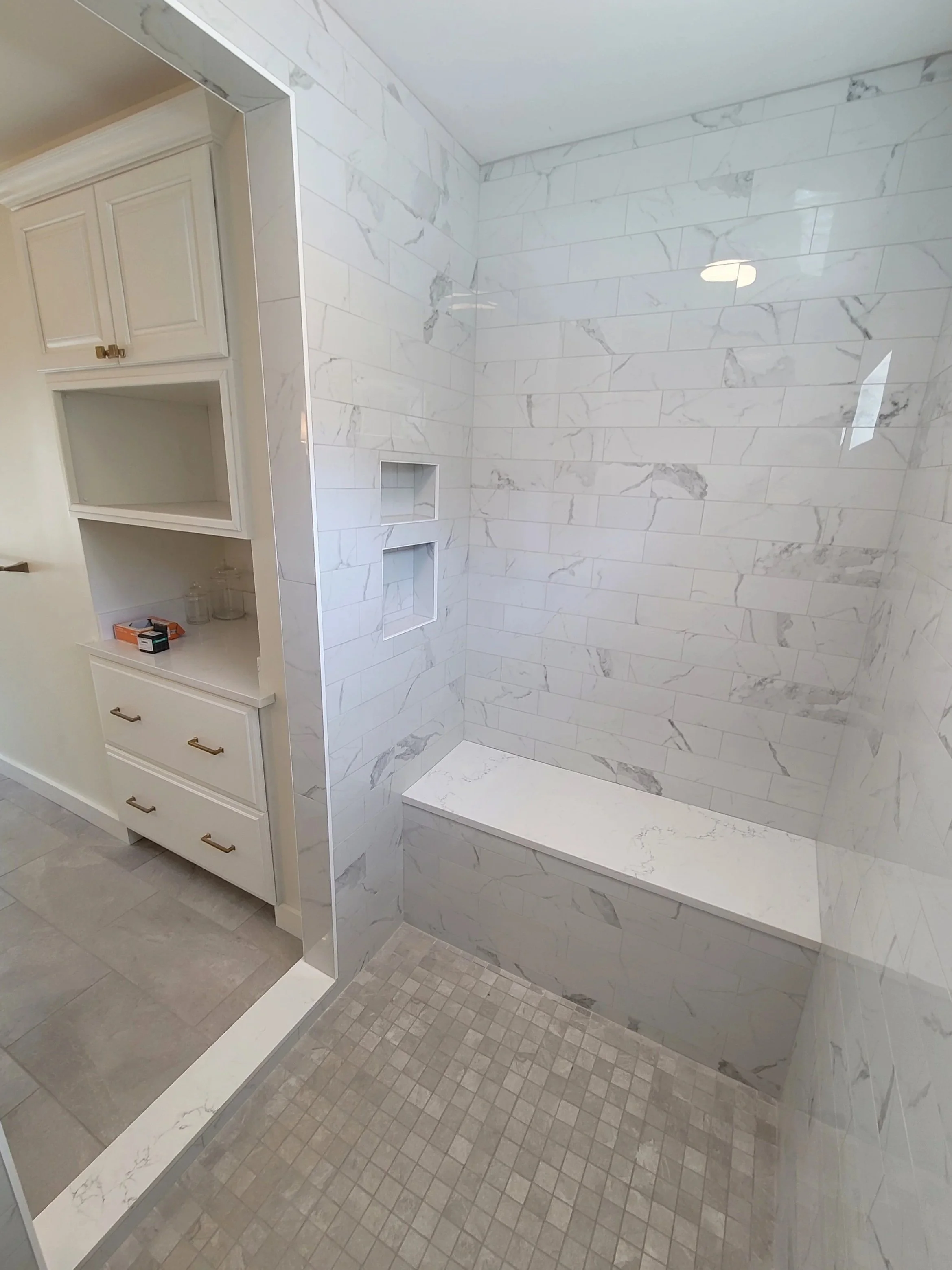 A modern bathroom shower area with white marble tiles, built-in niches for toiletries, and a partial view of a white bathroom shelving unit with decorative jars and boxes.