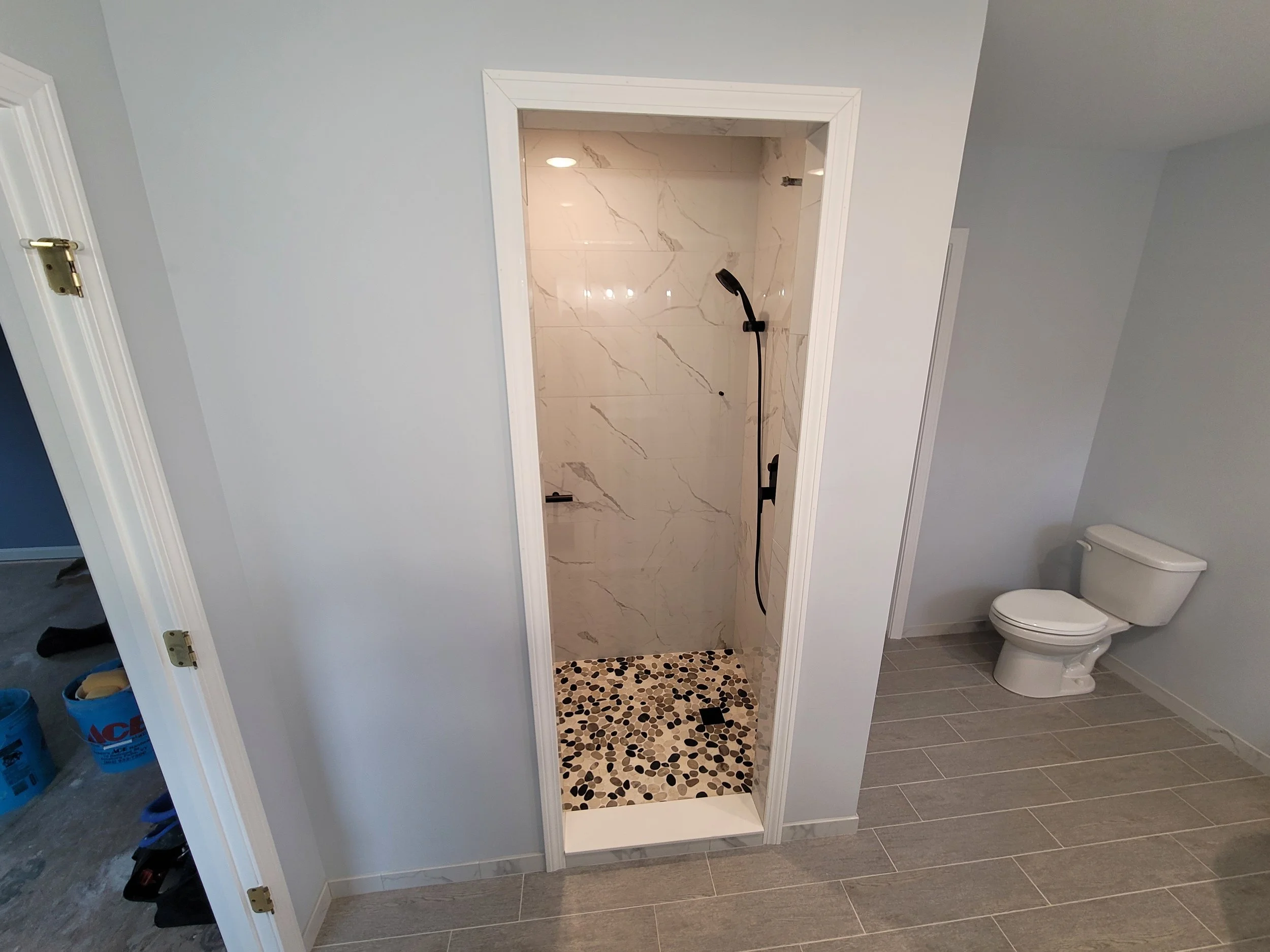 A walk-in shower with marble wall tiles, pebble floor tiles, and black shower fixtures, situated in a bathroom with gray tiled flooring and a toilet nearby.