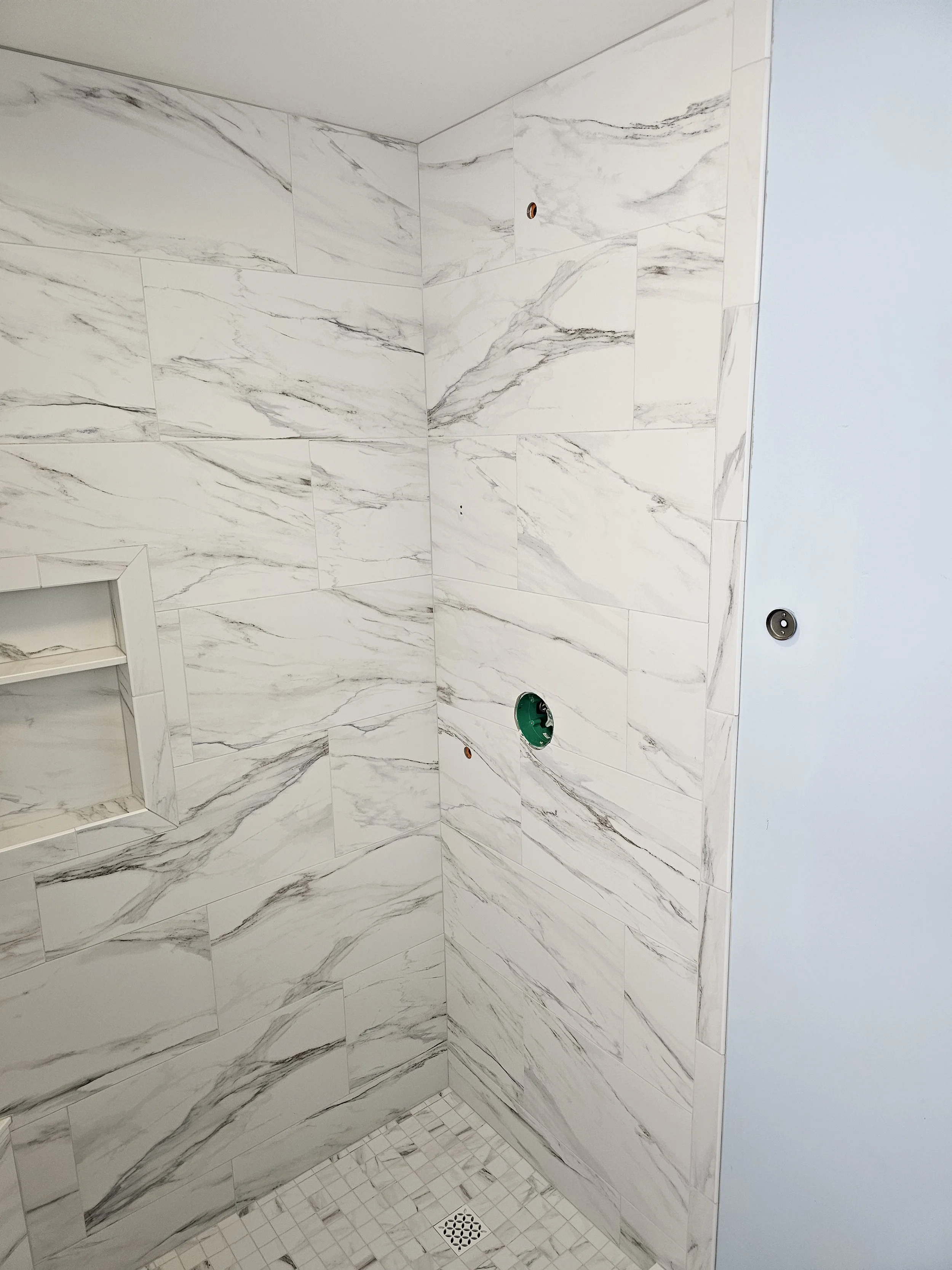 Construction of a tiled shower with marble-look tiles, a small built-in shelf, and a drain on the floor. The wall has exposed plumbing fixtures.