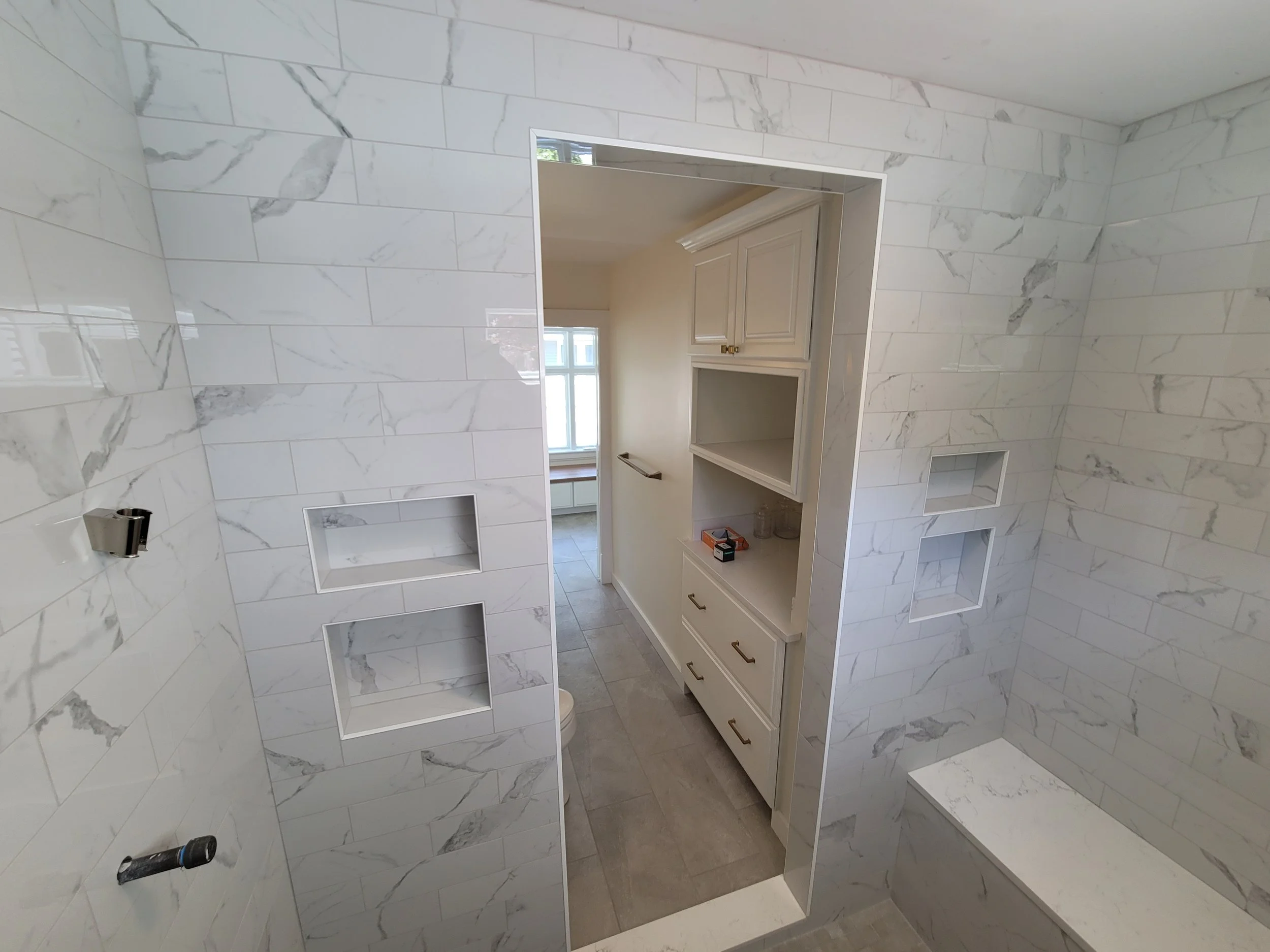 A bathroom with a wall covered in white marble tiles with gray veining. There is an open cabinet with drawers and shelves, a window letting in natural light, and a partially visible mirror. The floor has gray tiles and a small black object, possibly a flashlight or a tool, is on the floor.