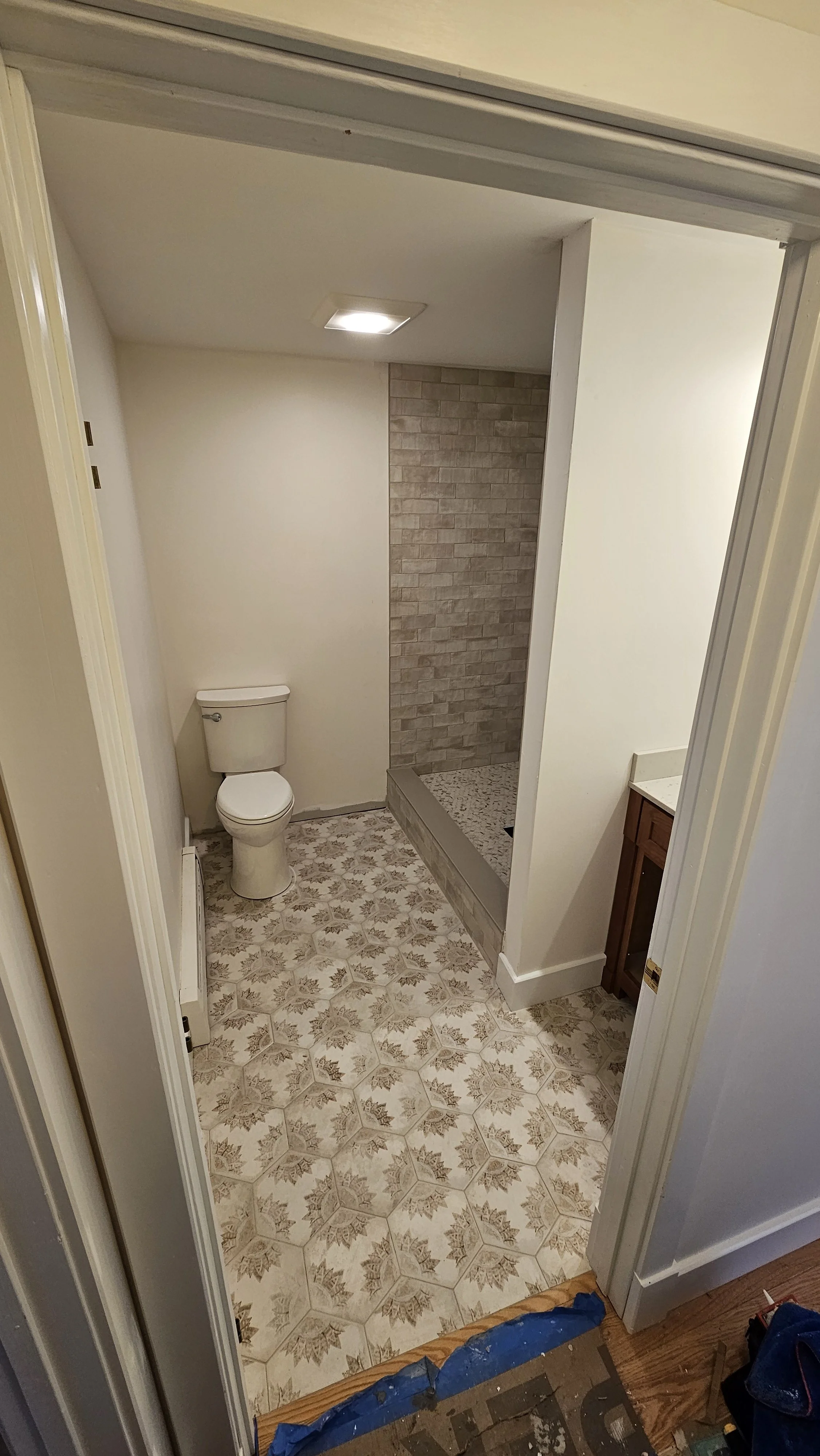 Bathroom with toilet, shower area with brick tile, patterned vinyl flooring, and wooden cabinet.