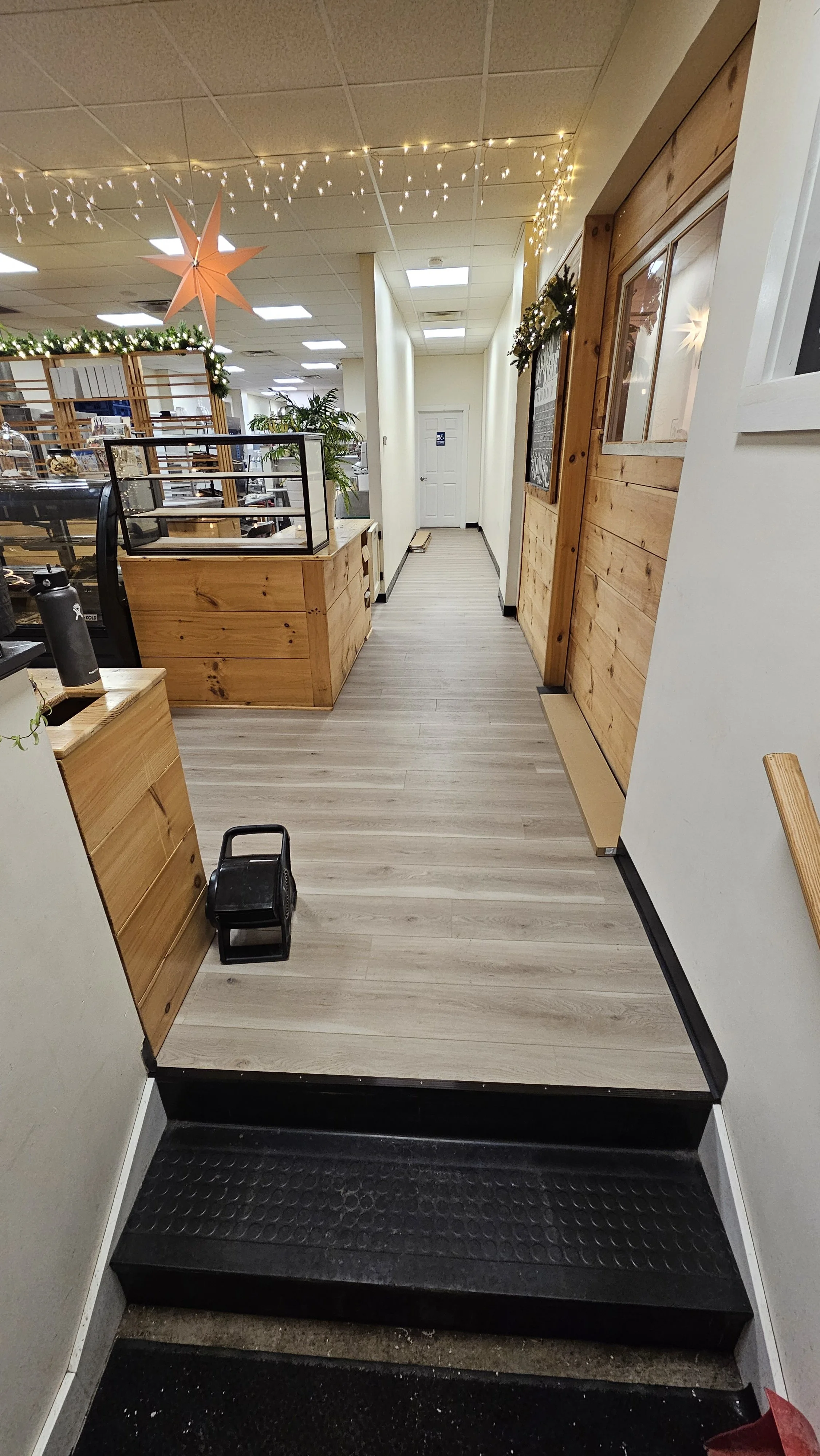 Interior view of a cafe or bakery with wooden accents, fairy lights, and a display case. A stairway with black steps leads up inside the establishment.