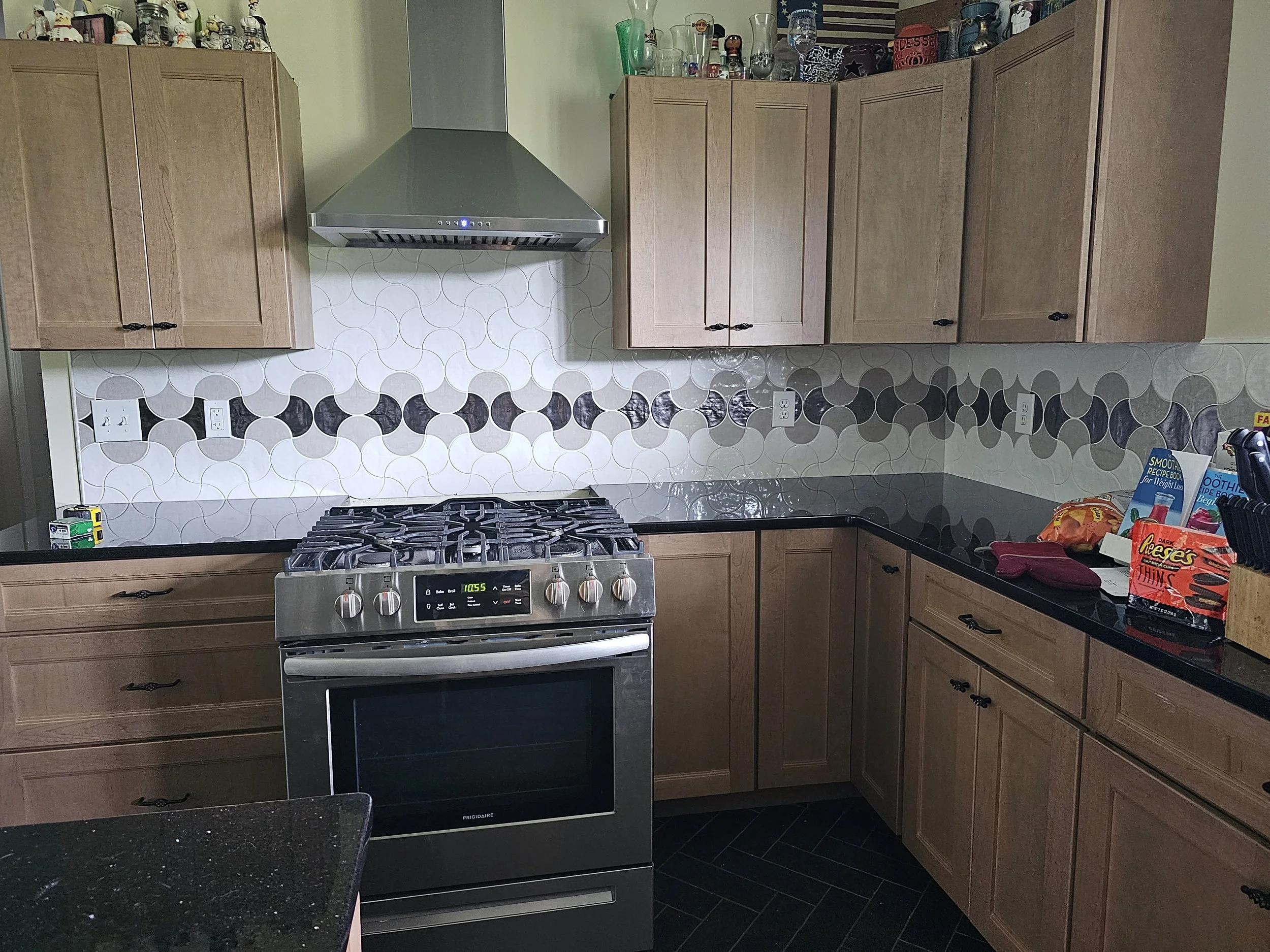 A kitchen with light wooden cabinets, a stainless steel stove and range hood, a patterned tile backsplash, black countertops, and various dishes and kitchen items on top of the cabinets.
