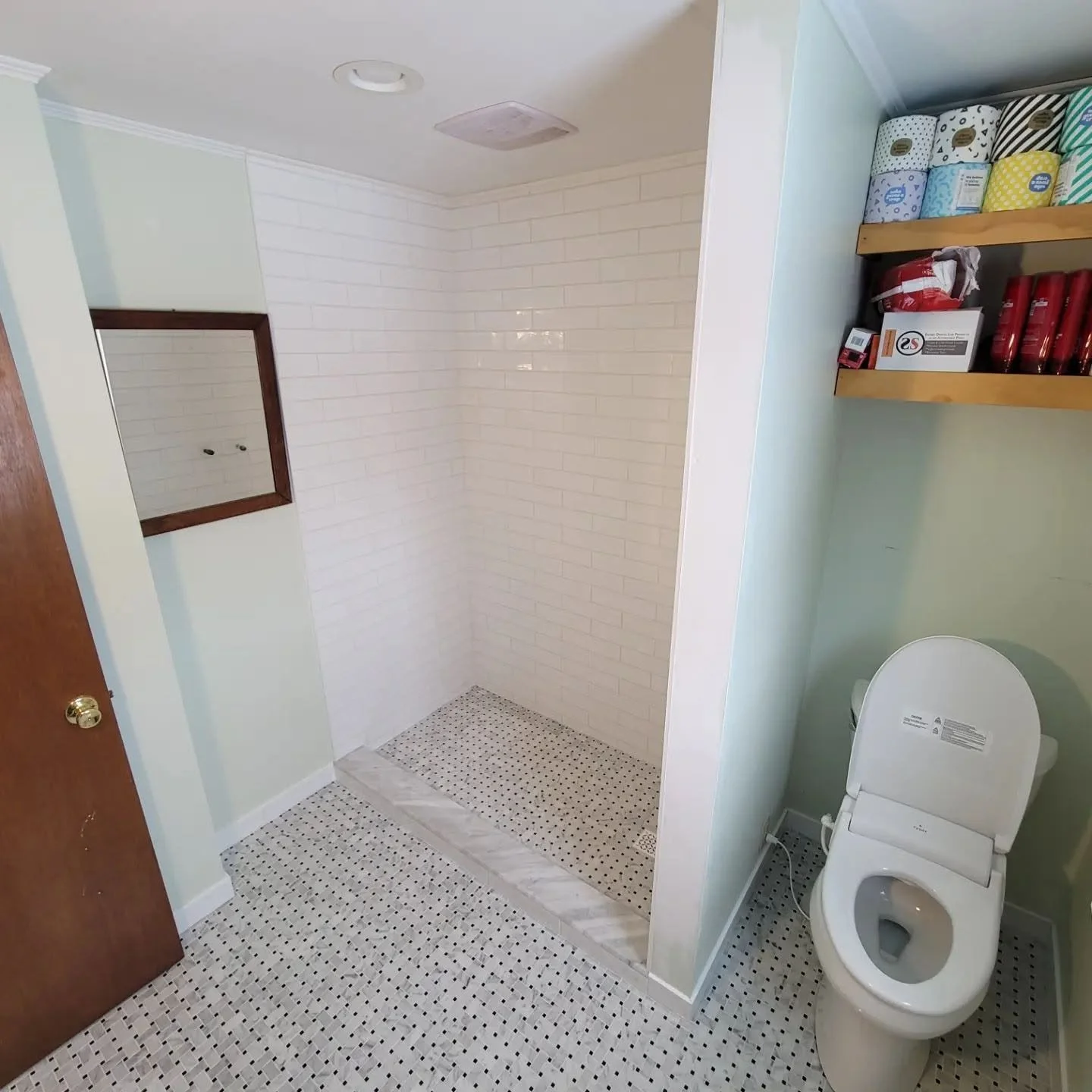 Bathroom with a toilet and an open shower area with white brick walls, small mosaic tile floor, a wooden shelf with paper towels and toilet paper rolls, and a mirror on the wall.