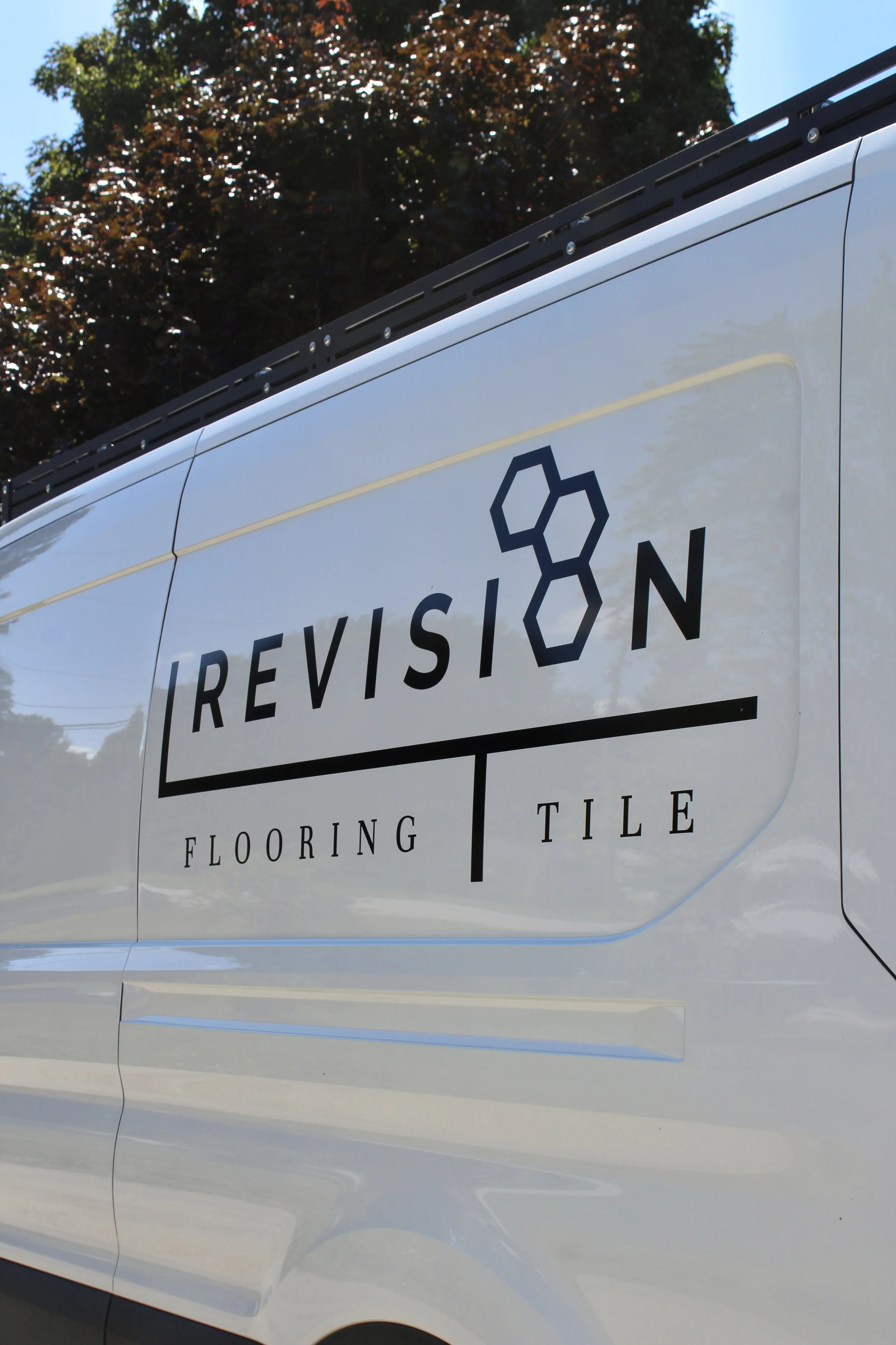 Close-up of a white van with a logo and text that reads 'REVISION' and 'FLOORING TILE' on the side. The background shows trees and a clear blue sky.