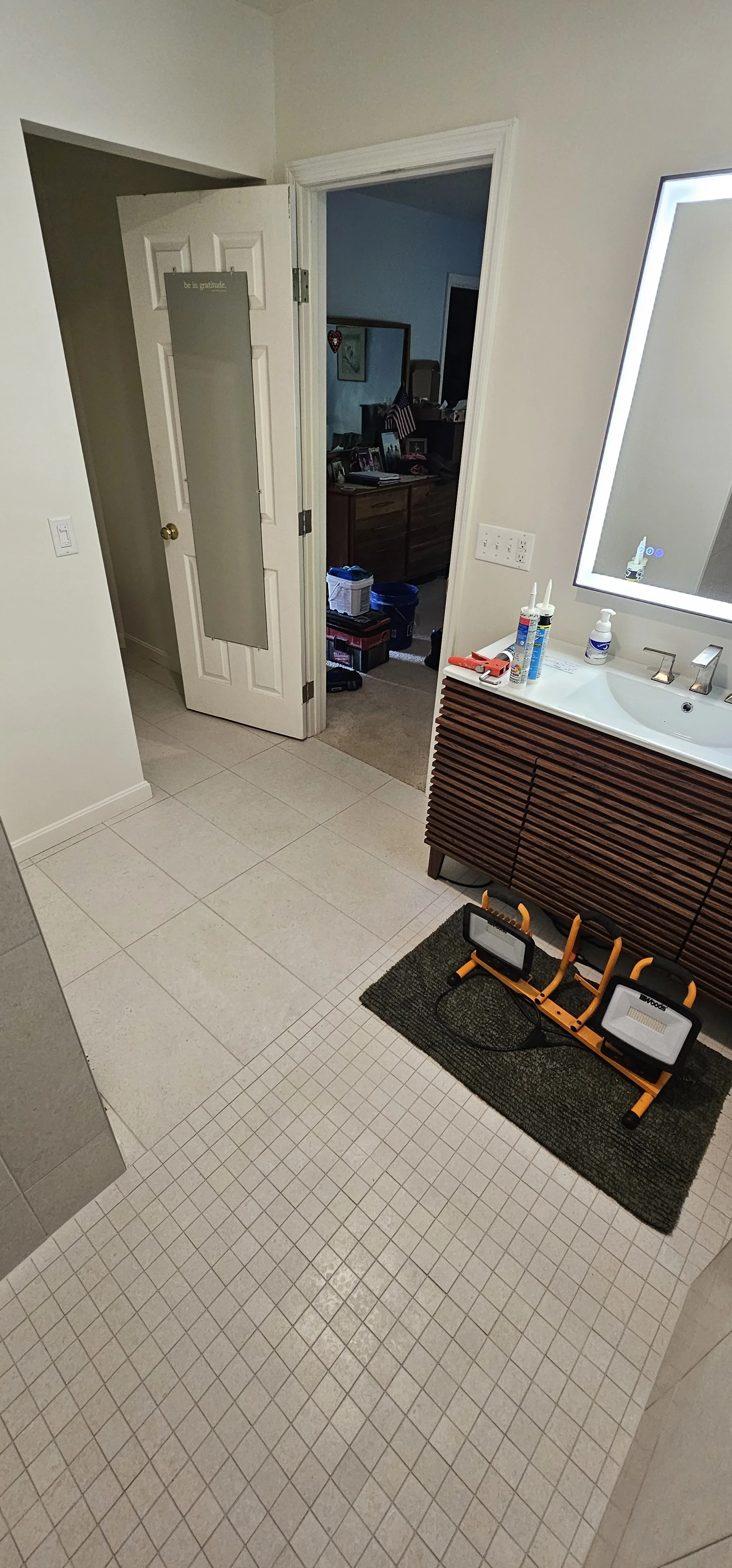 A bathroom with a textured tile floor, a wooden vanity with a mirror, and a small exercise bike on a mat. The door leading to another room is open, revealing clutter and furniture.