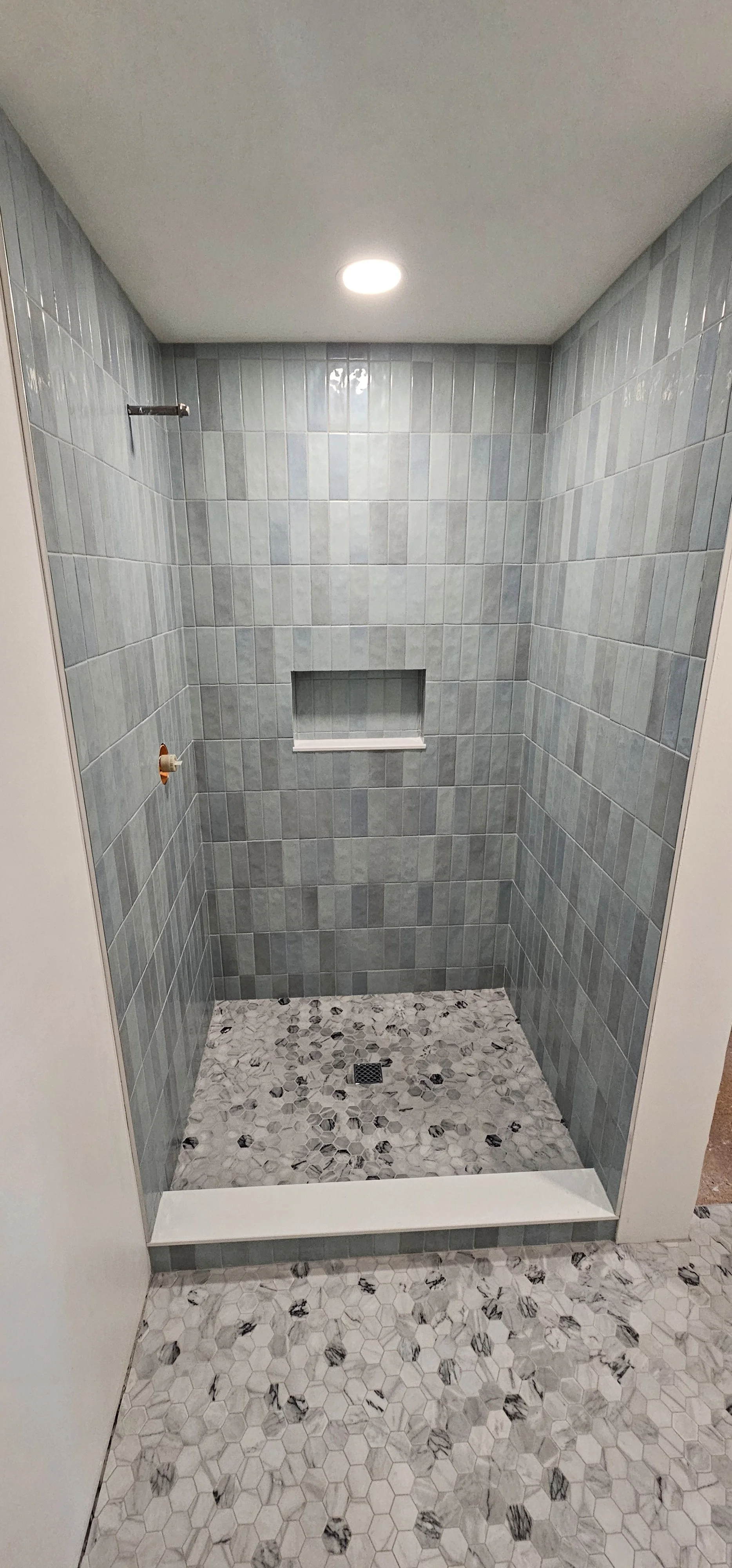 A walk-in shower with green rectangular tiles on the walls, a built-in shelf, and a mosaic tiling floor with hexagonal and pebble patterns.