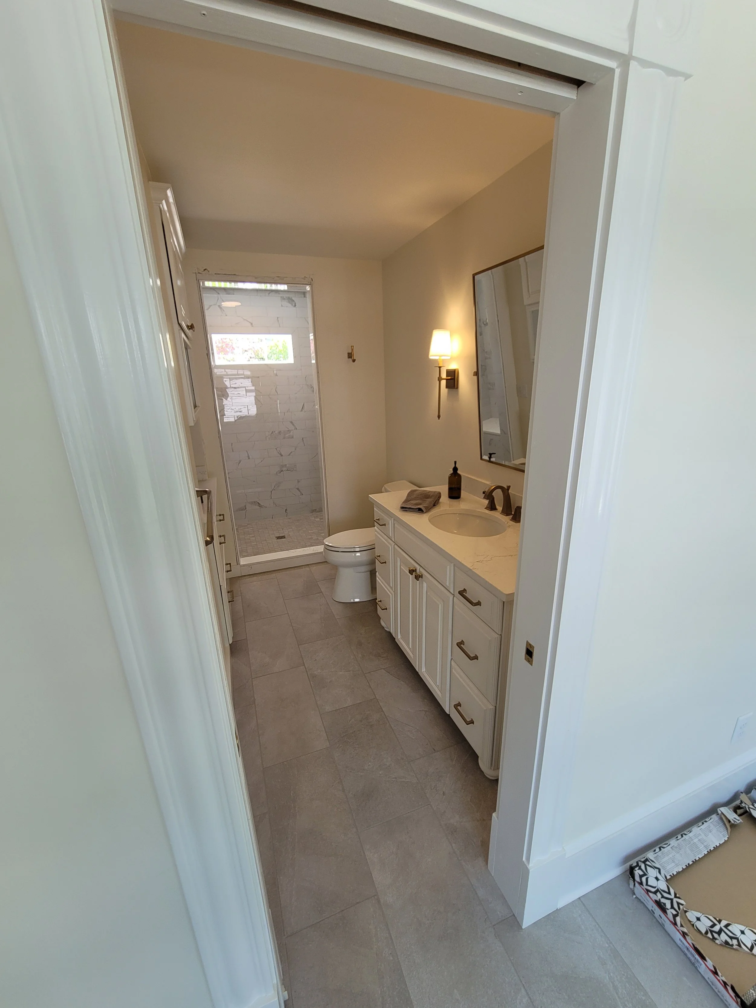 A small bathroom viewed through a doorway; features a white vanity with a marble top, a mirror, a wall-mounted light, a toilet, and a shower with a glass door and marble tiles.
