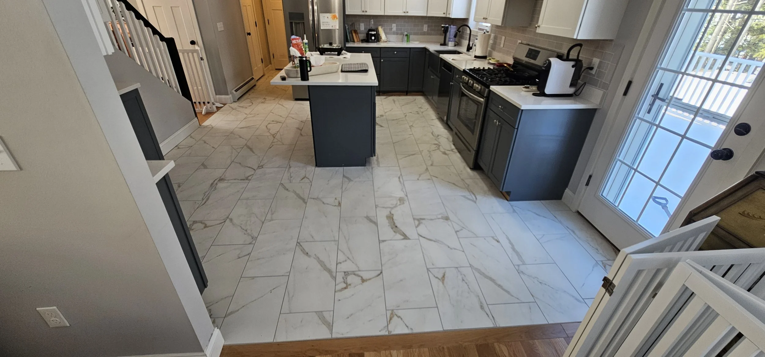 A kitchen with white and gray cabinets, a marble tiled floor, a white countertop island with objects on it, and a glass door leading outside.