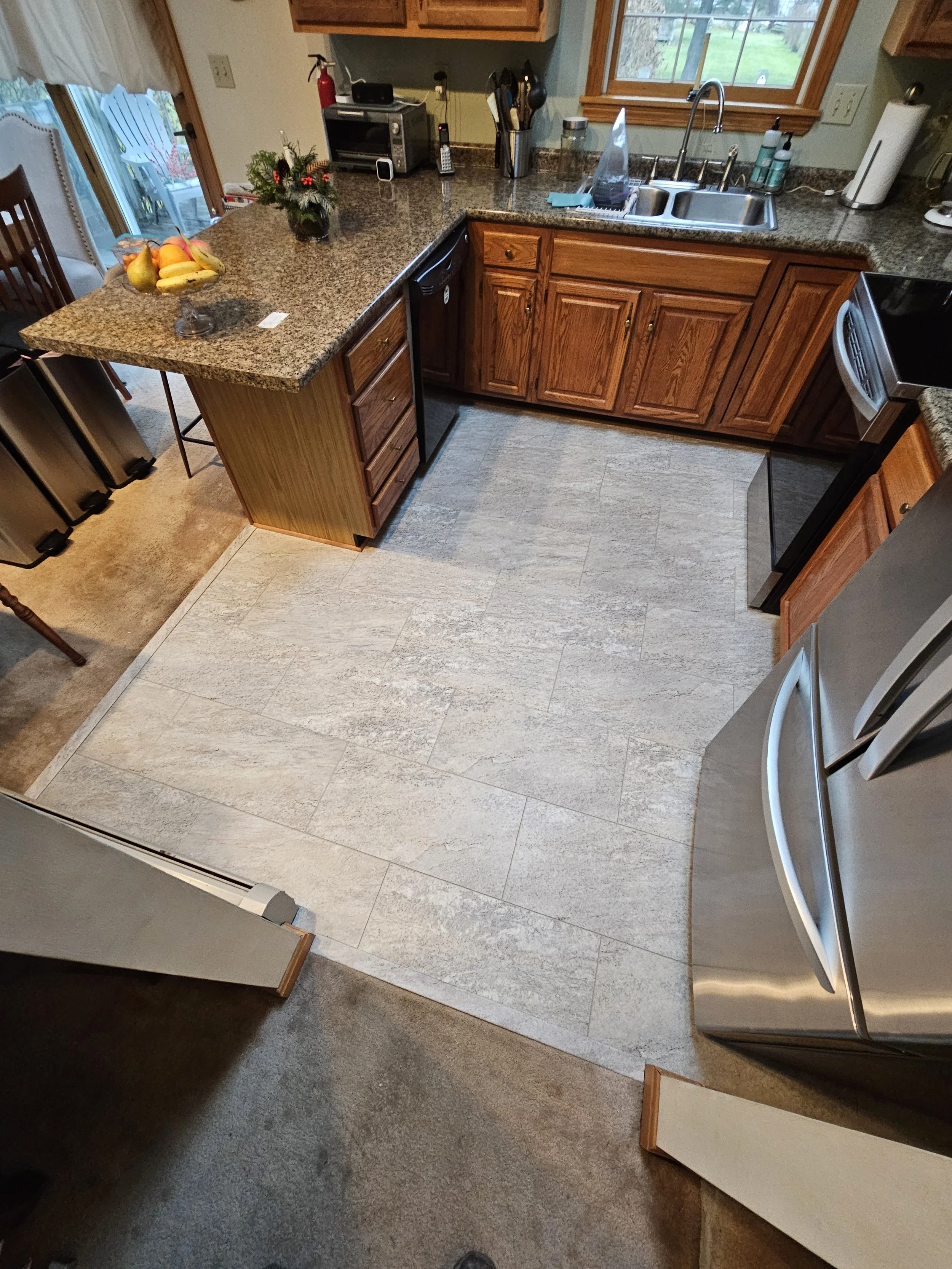 A kitchen with wooden cabinets and granite countertops, a stainless steel sink, a window above the sink, a black dishwasher, a fruit bowl with bananas and pears on the left counter, a small plant, a toaster oven, a cordless phone, and some utensils o