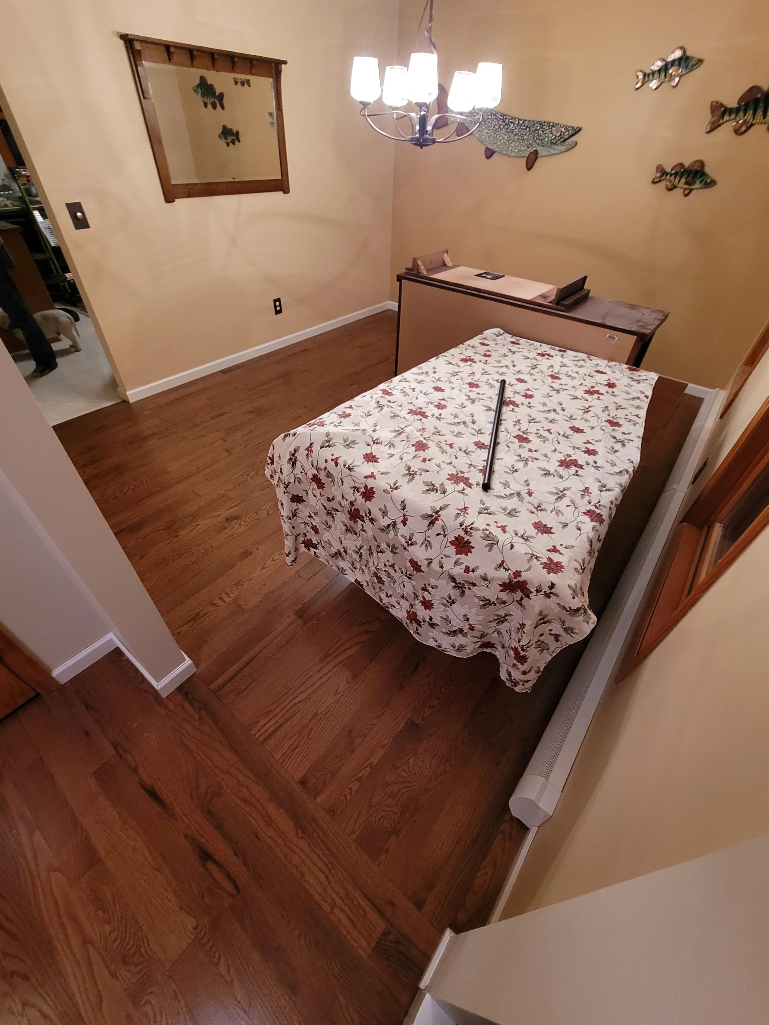 A small bedroom with a wooden floor, a bed with a holiday-themed bedspread, and a corner desk. Decor includes fish wall art and a ceiling chandelier with multiple lights.