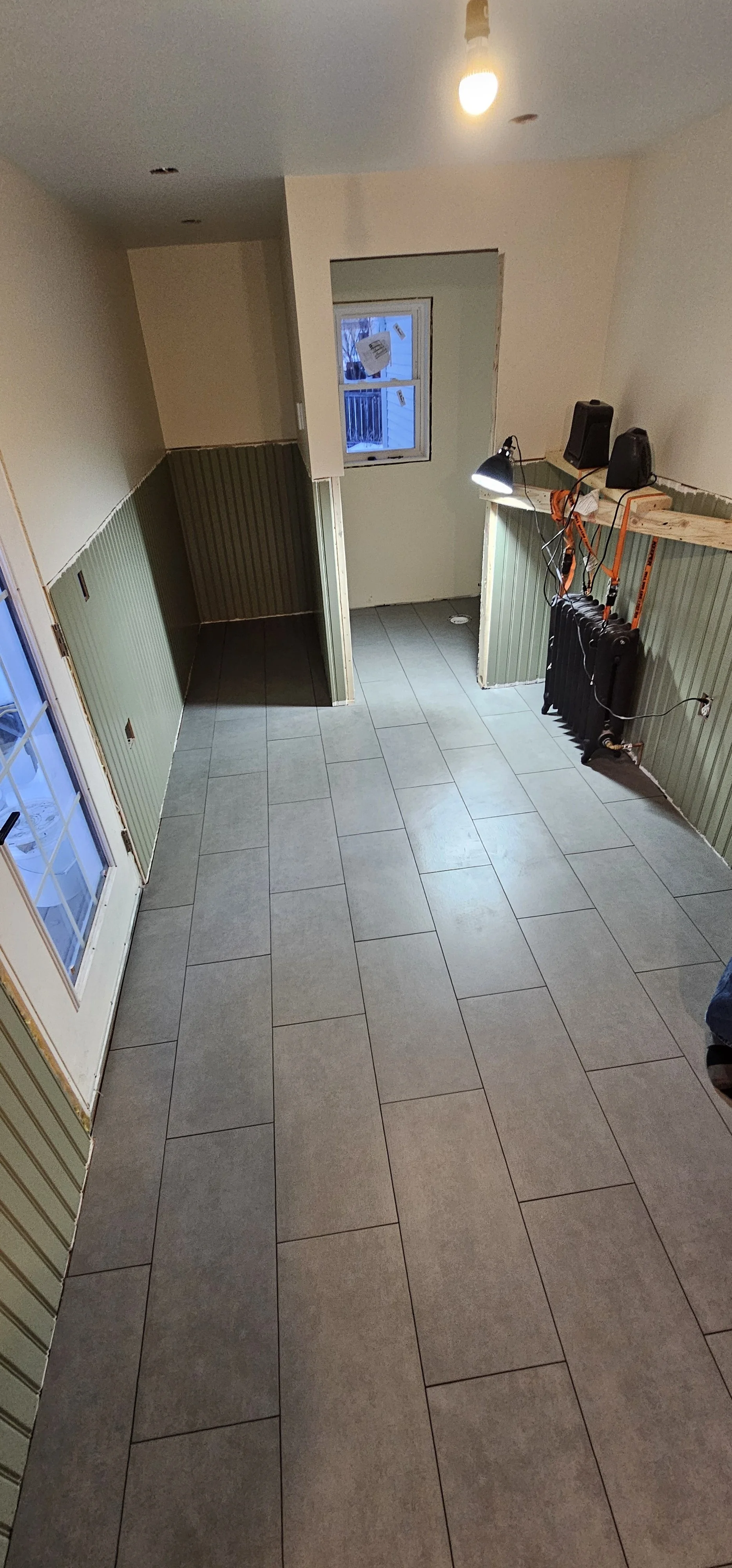 Room under construction or renovation with tiled floor, partially finished walls, a window, and a door, with construction tools and supplies on a shelf.