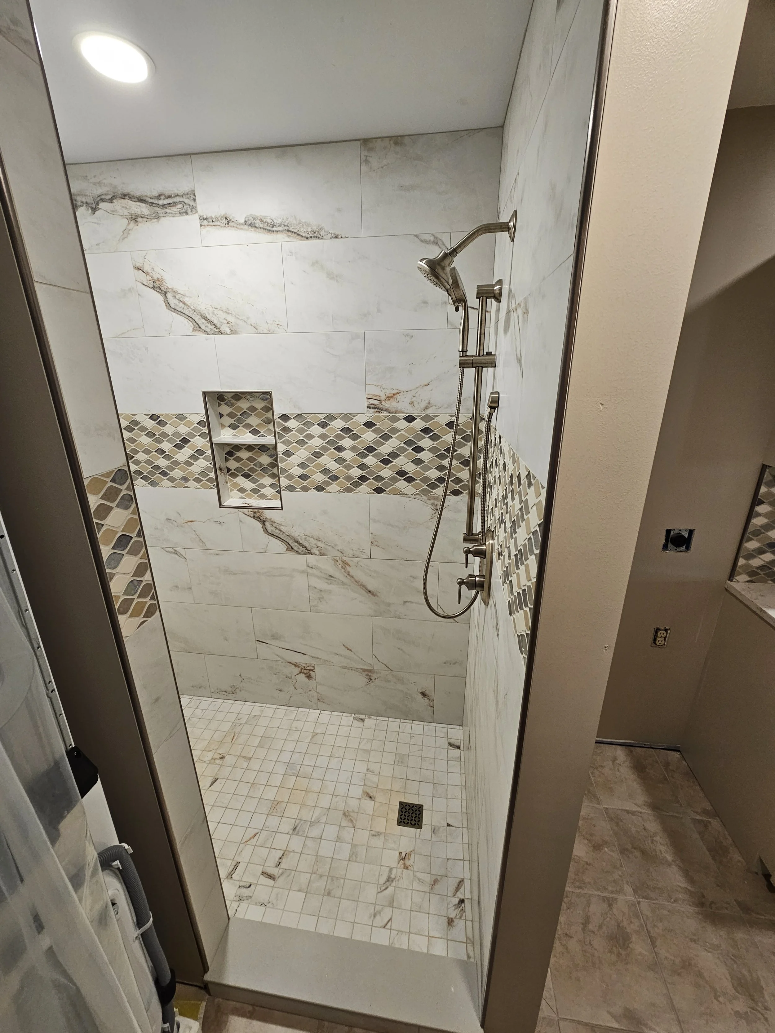 Shower with white and beige marble tiles, a built-in niche, a handheld showerhead, and beige floor tiles.