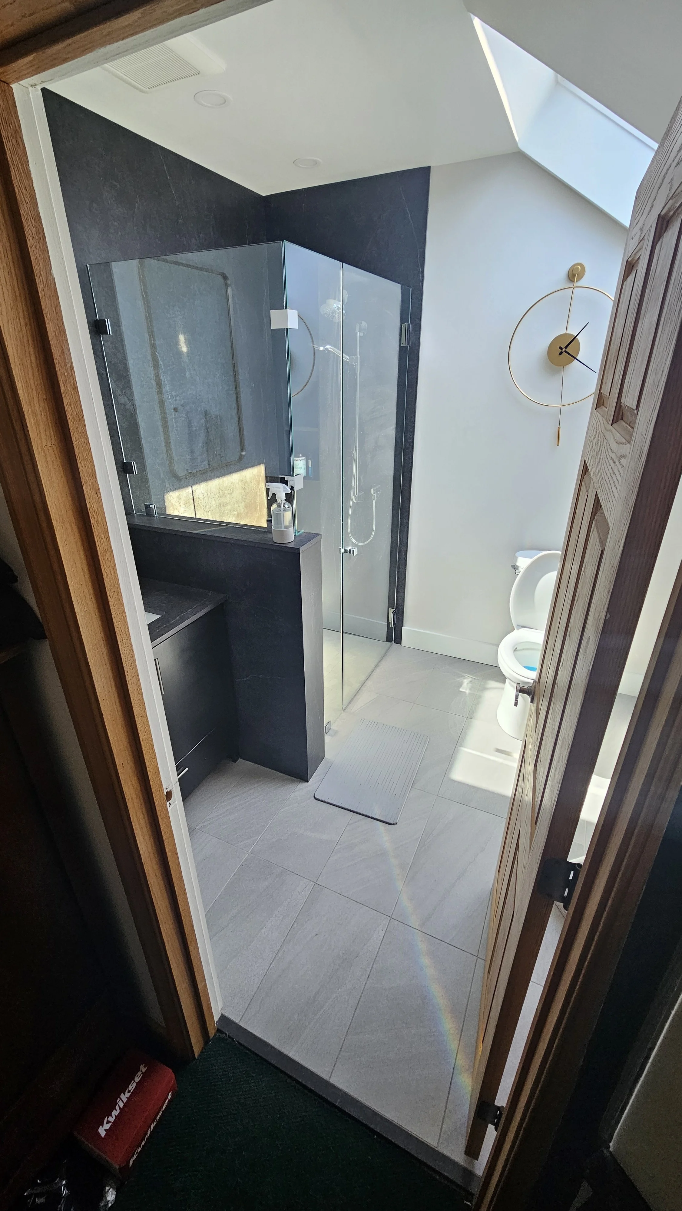 A modern bathroom with a glass shower, a wall clock, a toilet, a vanity with a spray bottle, a skylight on the ceiling, and a white bath mat on the tiled floor.