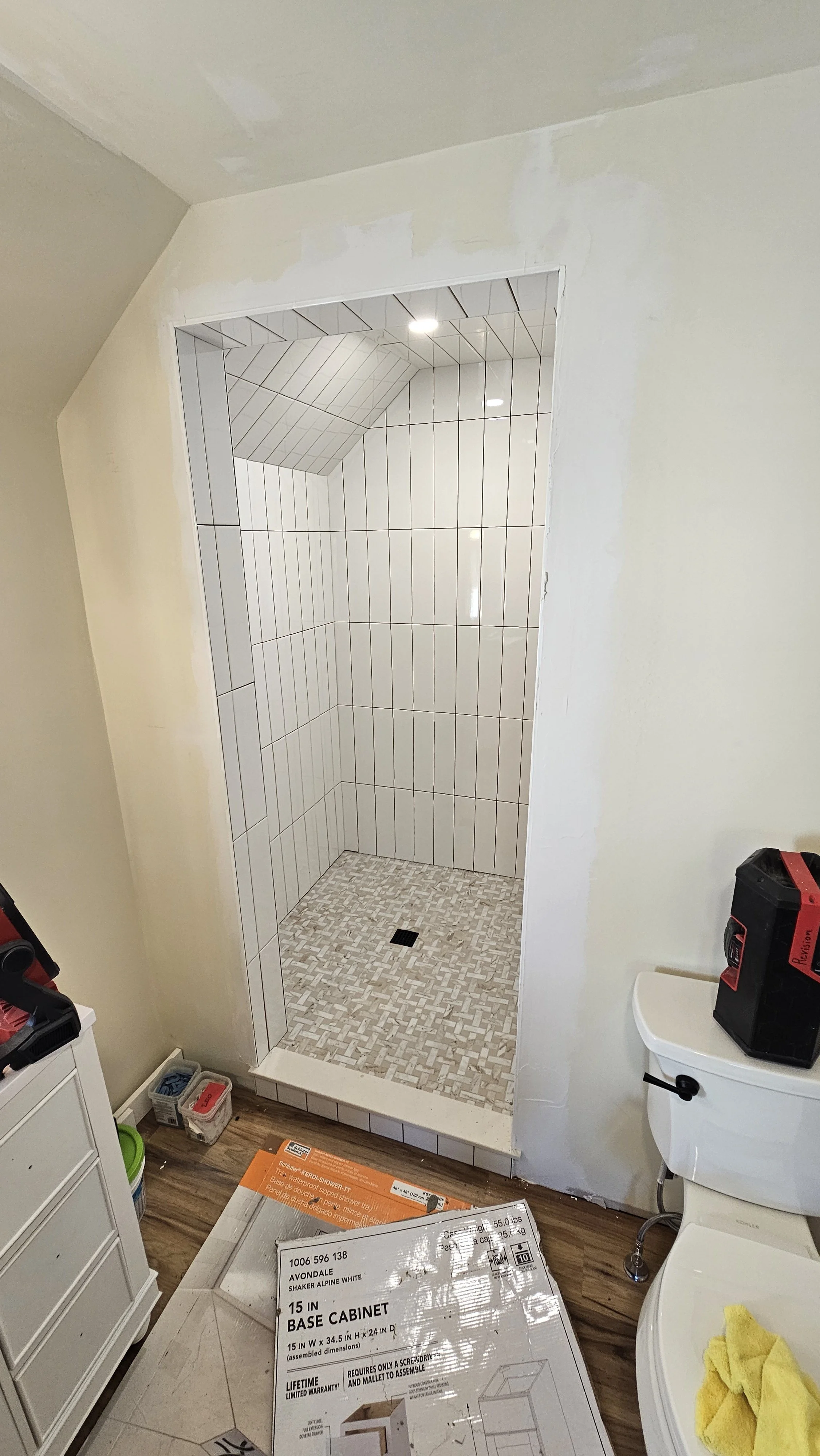 Bathroom shower with white tile walls, small mosaic tile floor, and a drain in the center. Construction tools and supplies are present, indicating ongoing installation or renovation.