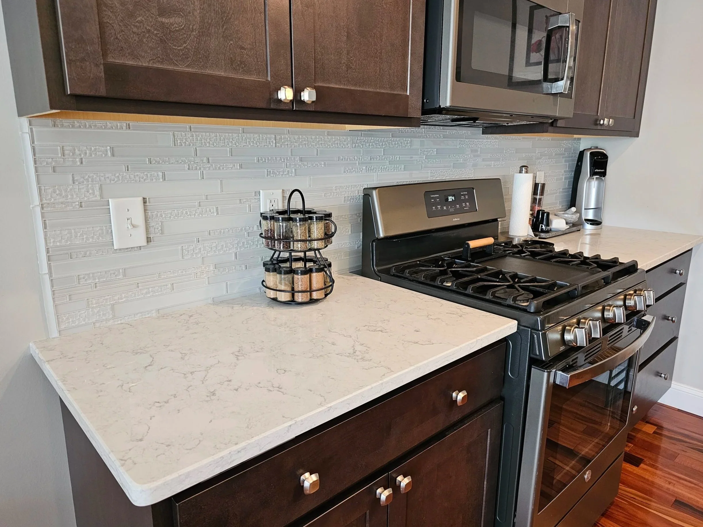 Kitchen countertop with a spice rack holding various spices, a microwave, a paper towel roll, a coffee maker, and a stove with oven. Dark wood cabinets and a textured white backsplash.