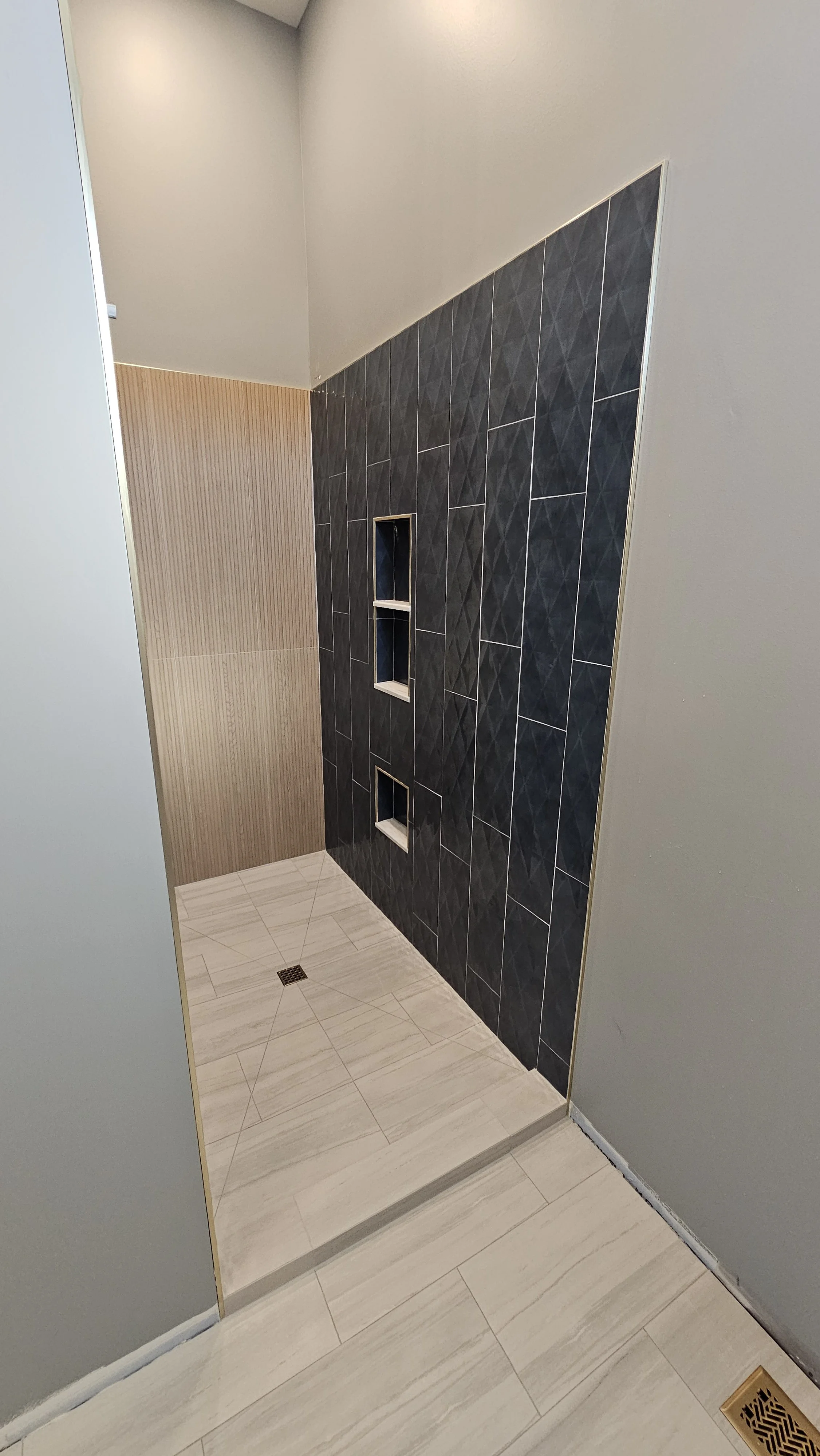 A modern shower area with black textured tiles on the wall, beige wood-like wall covering, and light-colored floor tiles, with small built-in shelving niches in the wall.