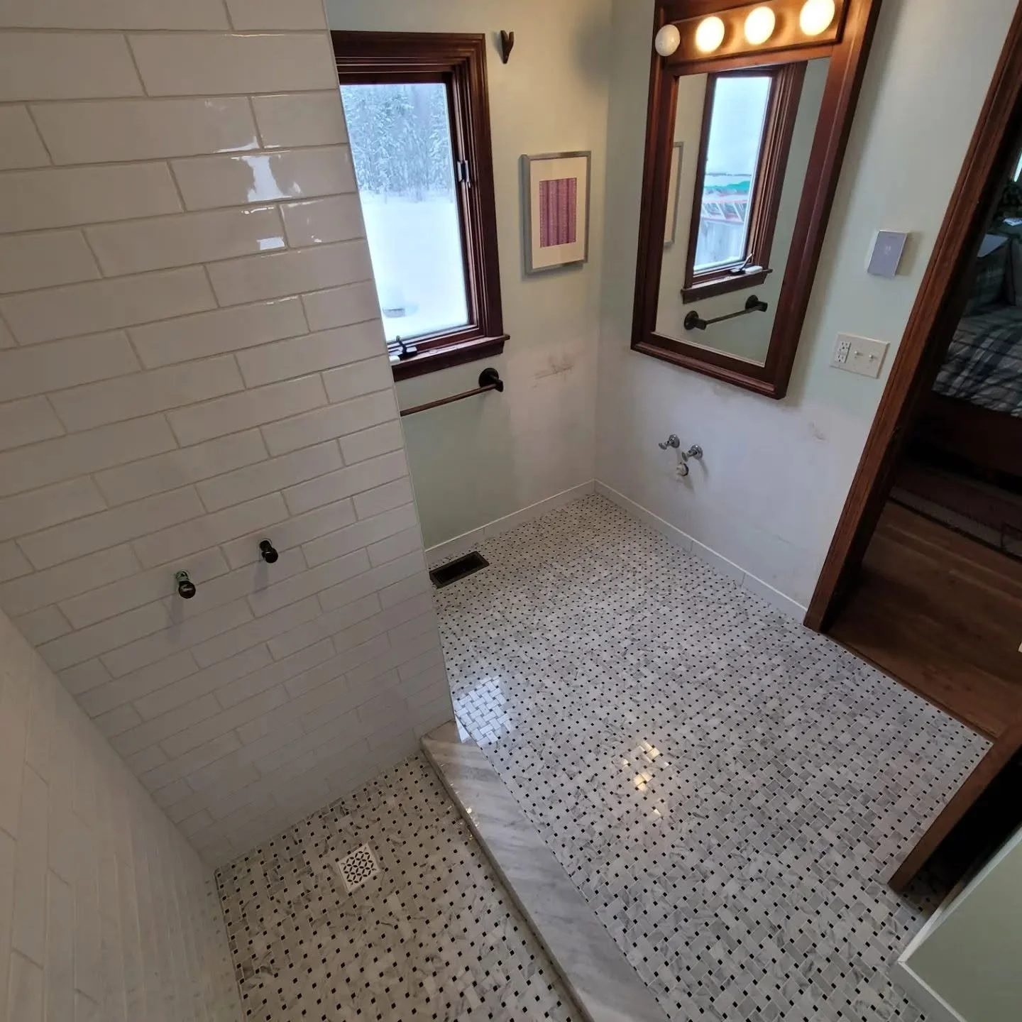 Empty bathroom with white subway tile on the wall, patterned mosaic floor, and two small windows with wooden frames.