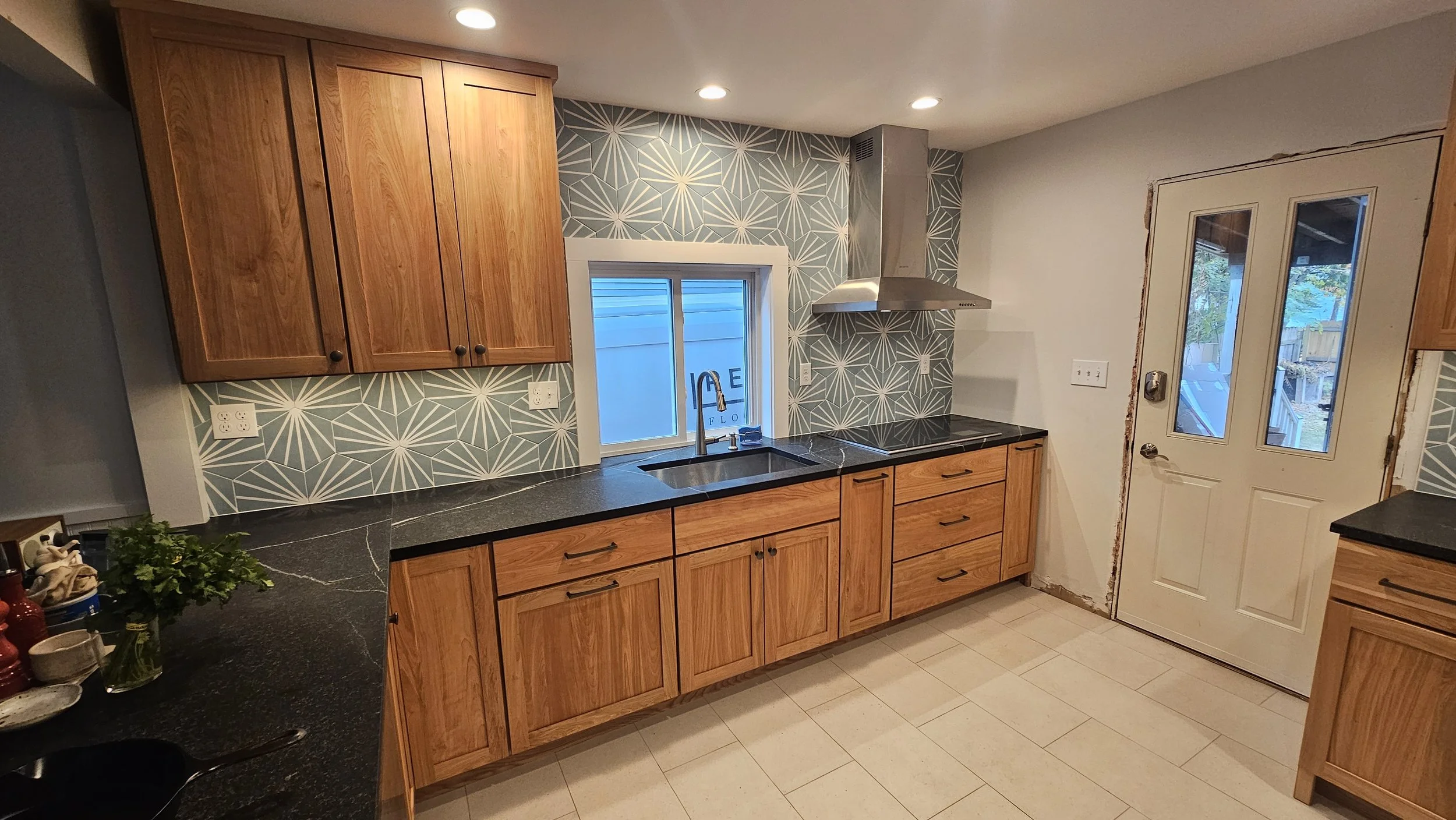 Modern kitchen with wooden cabinets, black marble countertops, geometric patterned blue and white backsplash, stainless steel range hood, small window over sink, white door with glass panels, tile flooring, and recessed ceiling lights.
