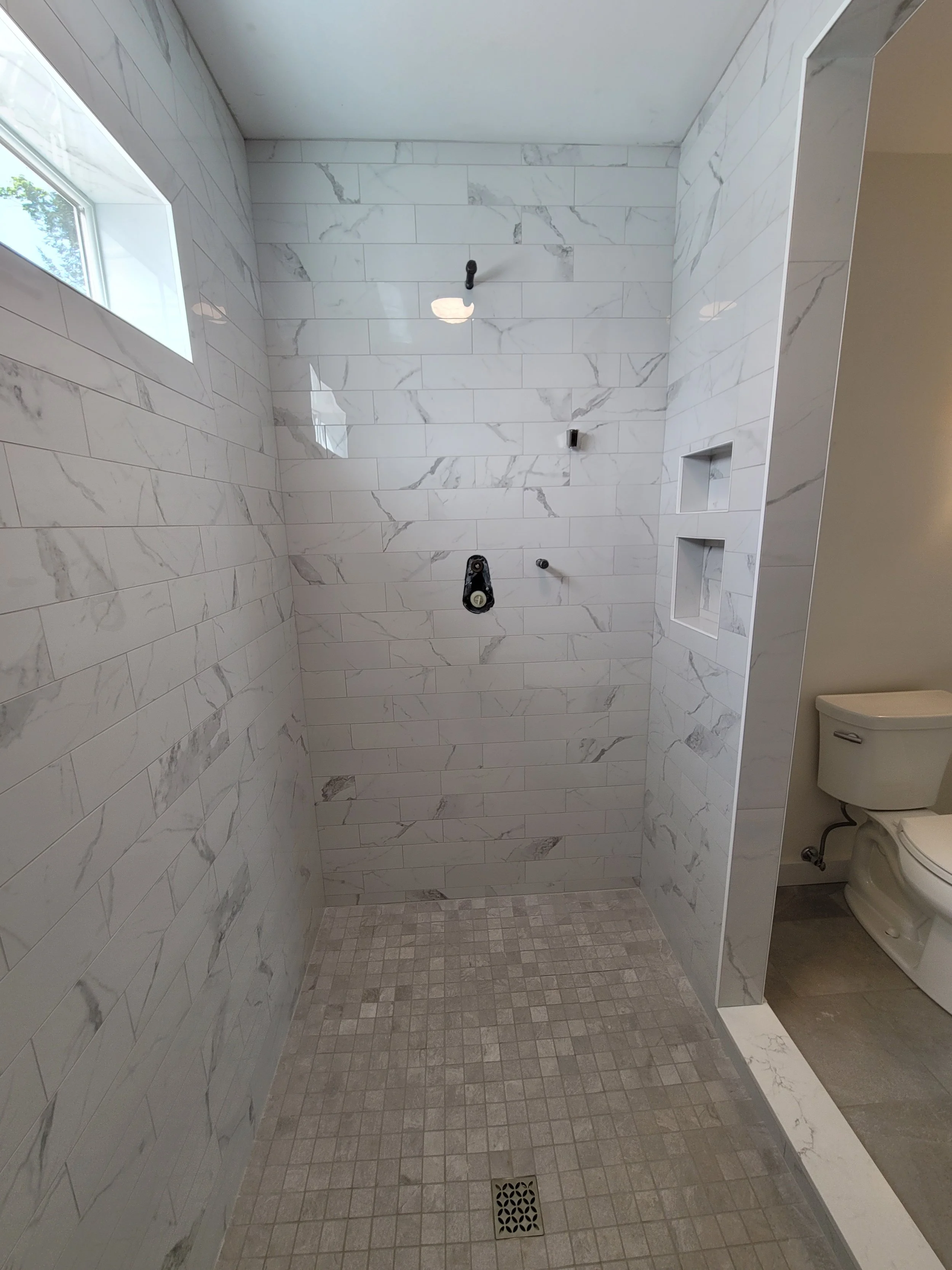 A walk-in shower with white marble wall tiles, a small window, and a square drain on the beige tiled floor. The shower is unfinished with fixtures not yet installed.