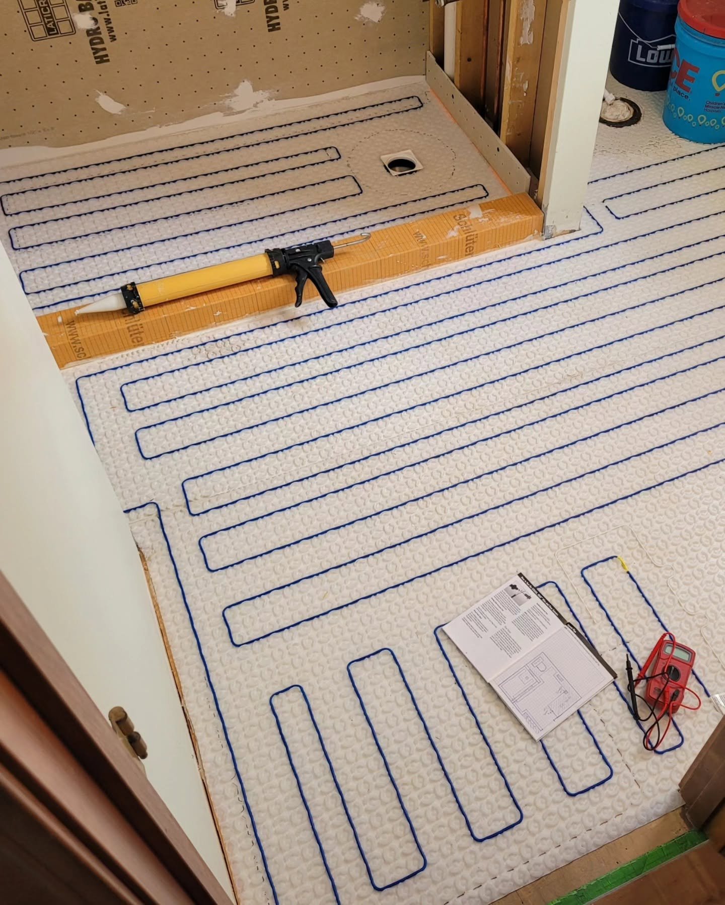Underfloor heating installation with blue tubing laid out in a pattern on the floor, construction tools, an instruction manual, and wiring equipment visible.