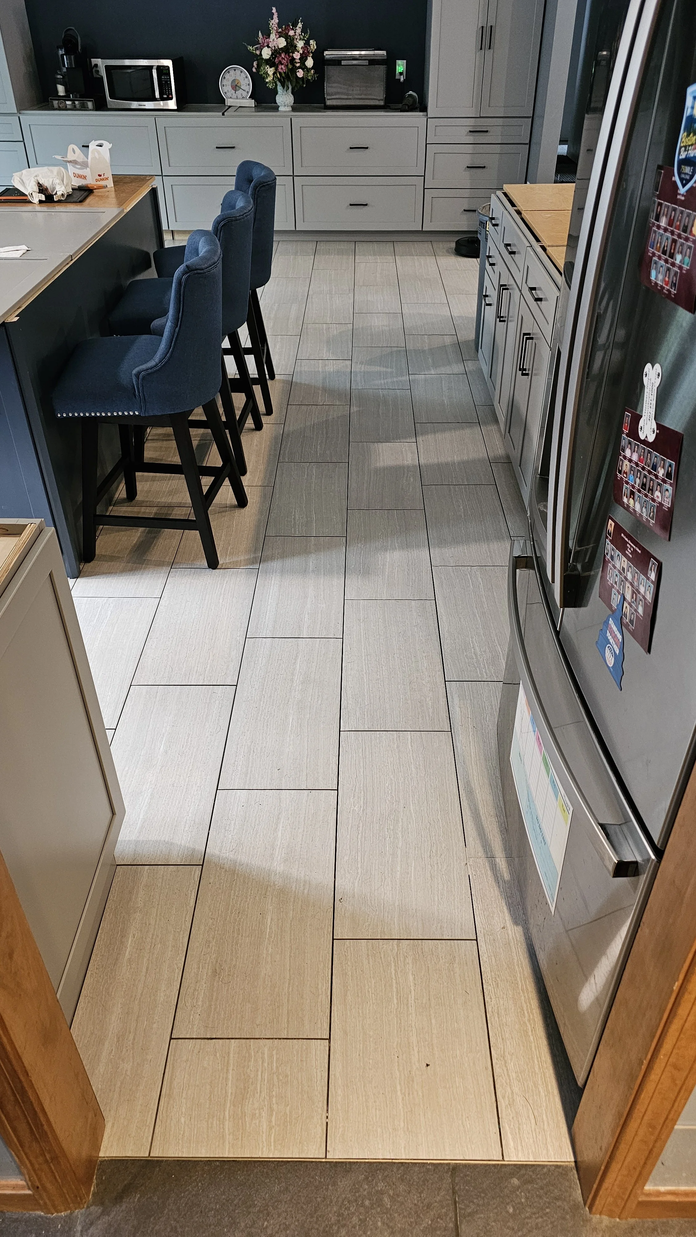 View of a modern kitchen floor with beige rectangular tiles, a stainless steel refrigerator, light gray cabinets, a breakfast bar with blue upholstered chairs, and various kitchen appliances including a microwave, toaster oven, and coffee machine on 