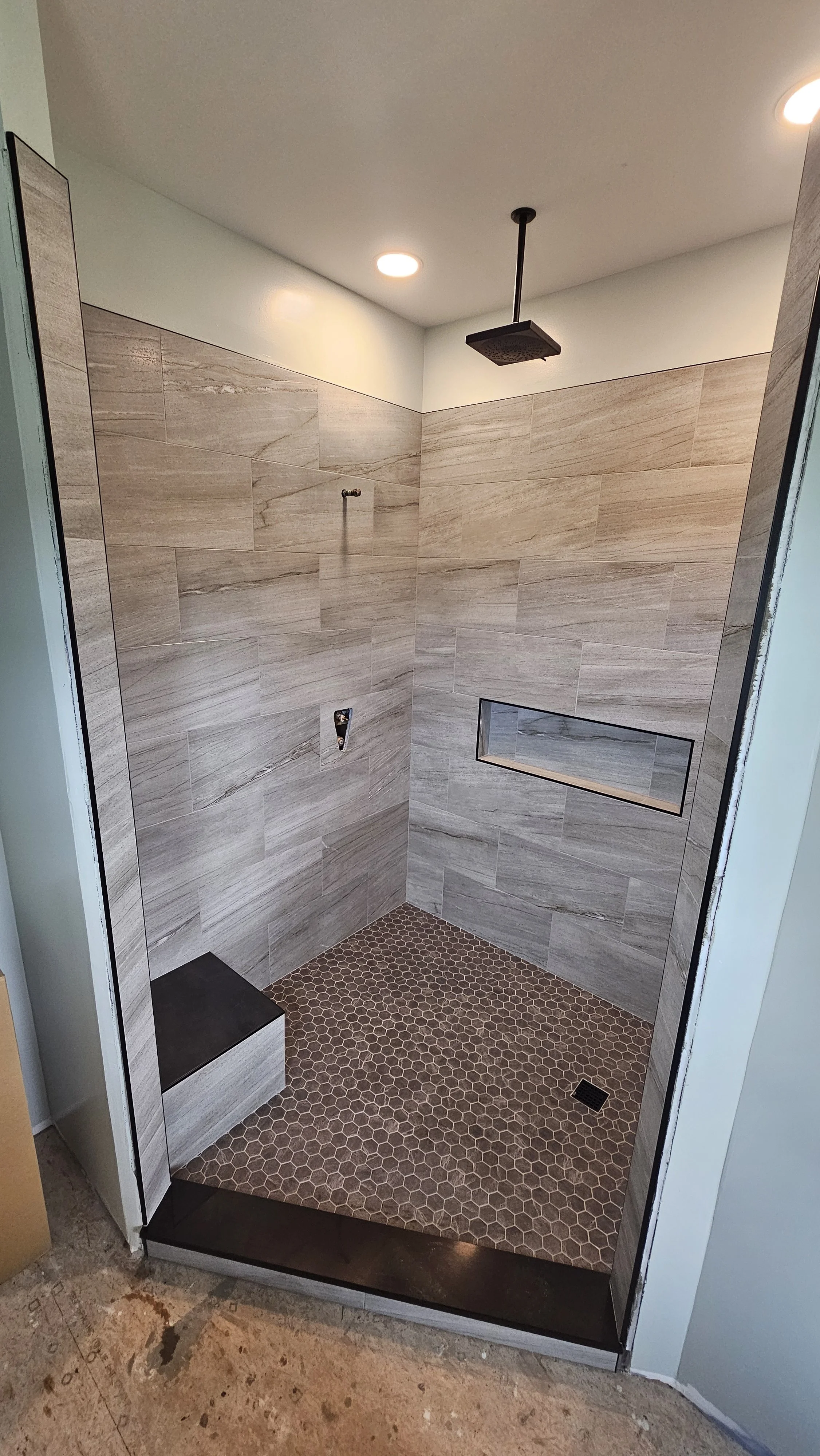 Modern shower with beige tiled walls, brown hexagonal tile floor, a built-in bench, a small rectangular niche in the wall, a ceiling rain shower, and recessed lighting.