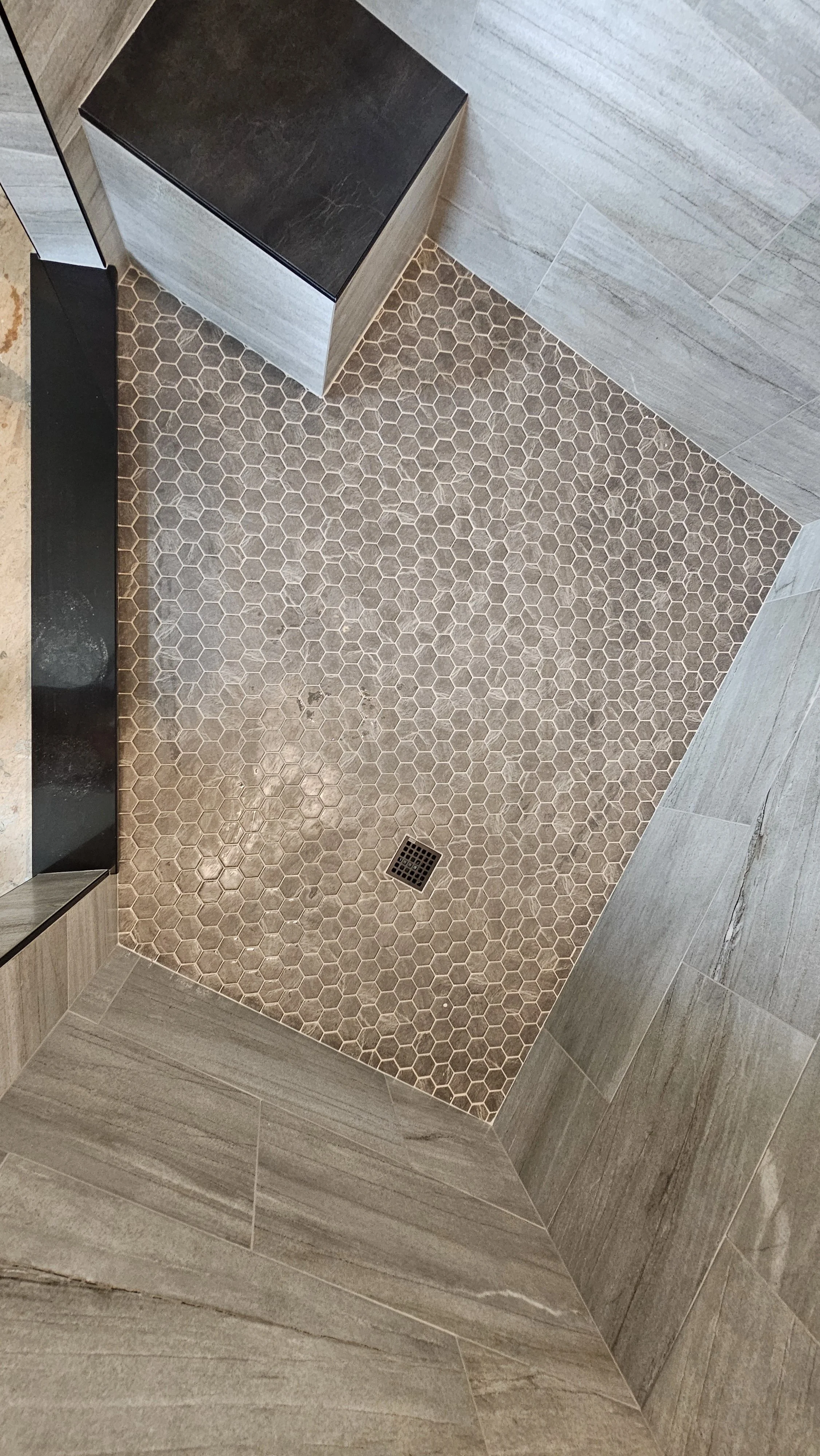 Shower floor with small hexagonal beige tiles, a square drain in the center, and surrounding walls covered in large rectangular gray tiles.