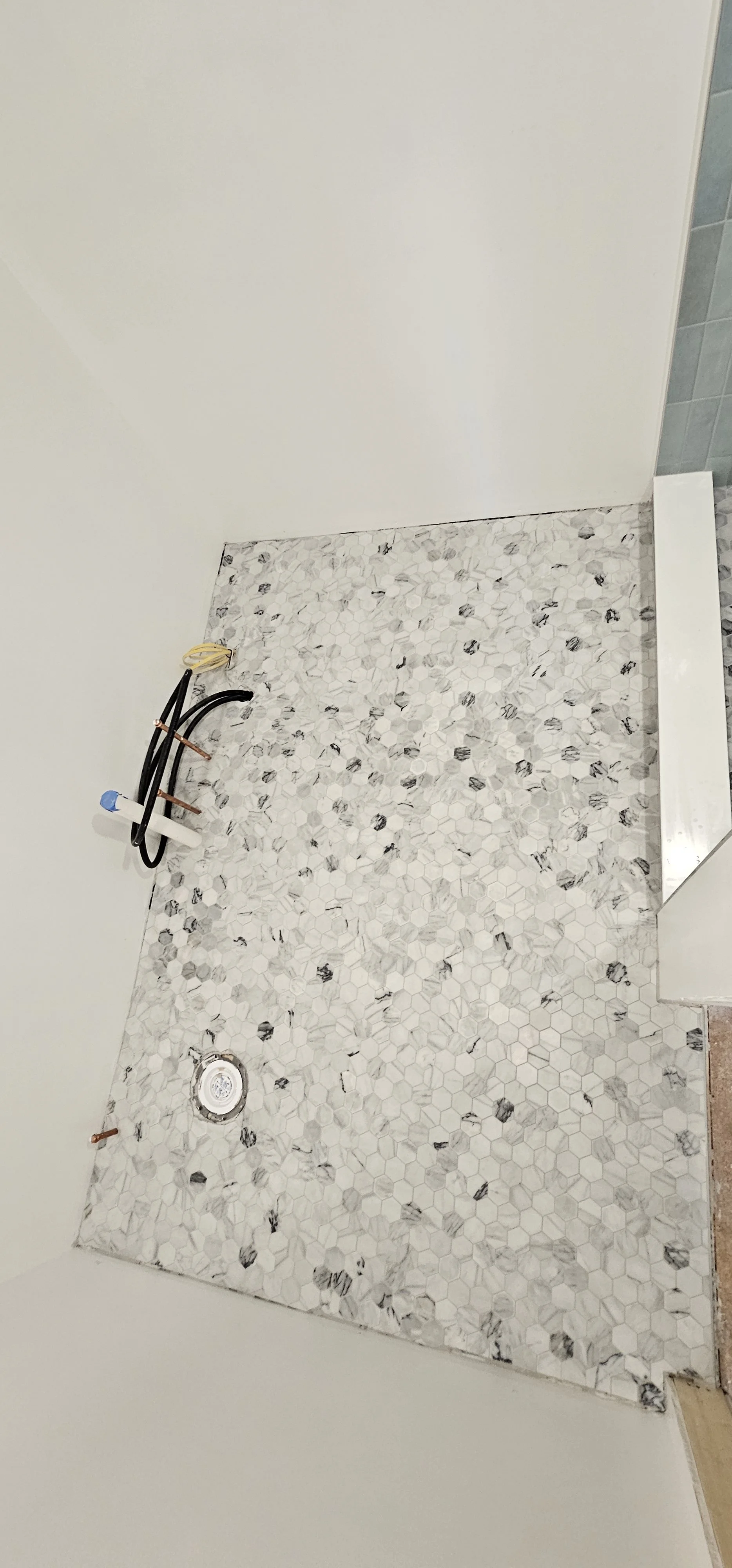 Bathroom floor with white hexagonal tiles with gray and black accents, plumbing pipes and drain, and partially installed vanity or cabinet.