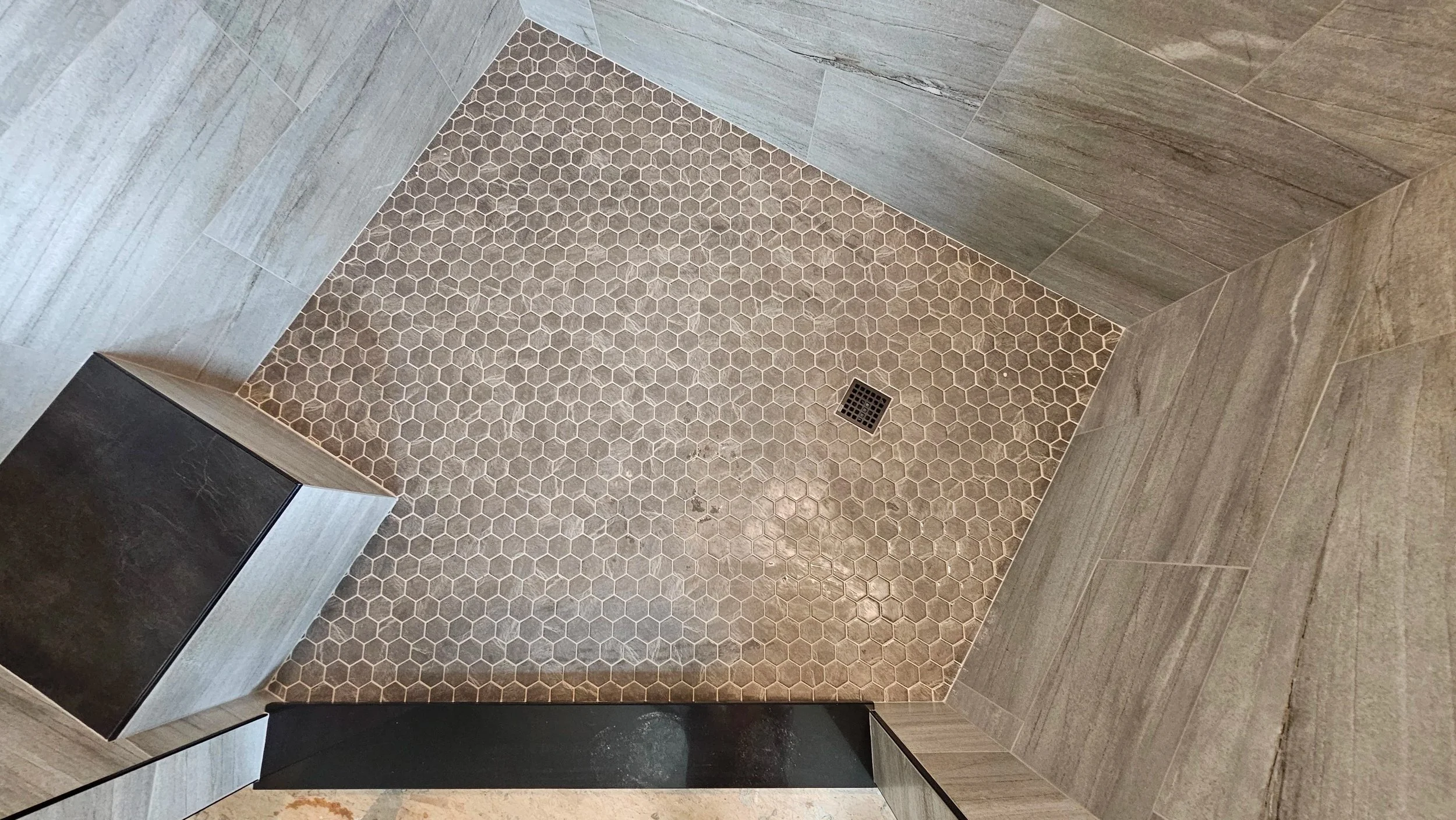 A close-up view of a shower area with beige hexagon tiles and a square drain, surrounded by gray rectangular wall tiles.