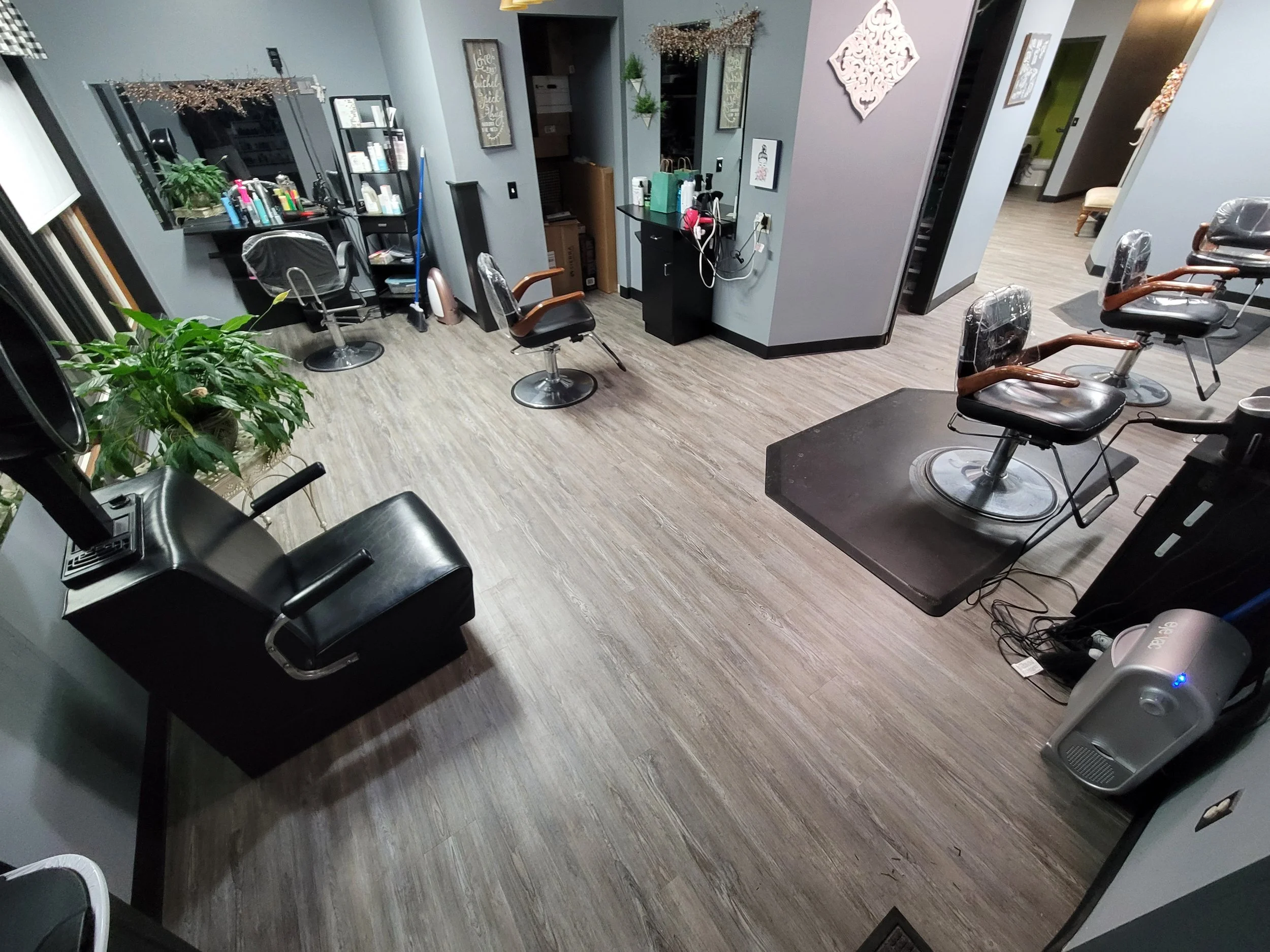 A hair salon with styling chairs, a mirror, and various hair products on a black shelf. The salon has wood flooring, decorative wall art, and a plant. A mirror reflects some of the chairs and equipment.