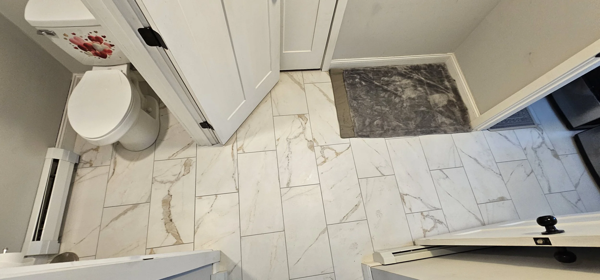 A bathroom with large white marble tiles on the floor, a toilet, a closed door, a gray mat, and a small heater or vent on the wall.