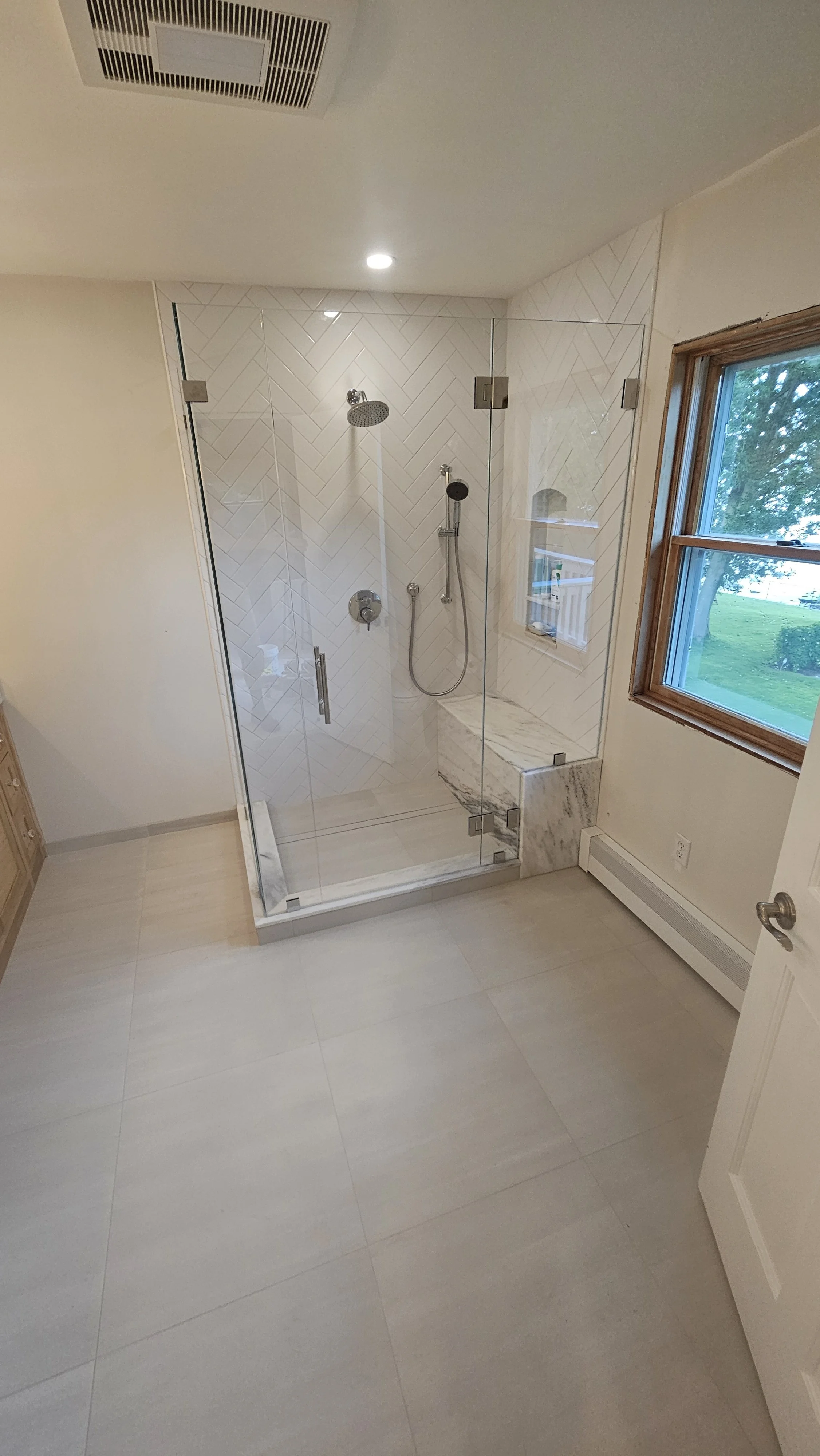 A modern bathroom with a glass shower enclosure, white herringbone tile on the shower wall, and a window showing green trees outside.