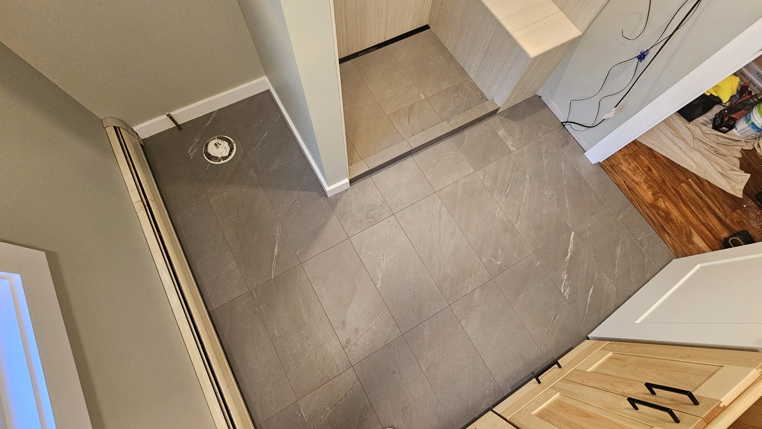 An unfinished kitchen or bathroom with dark gray tiled flooring, small steps, and cabinetry. Construction tools and materials are visible on the right side, with electrical wires hanging on the wall.
